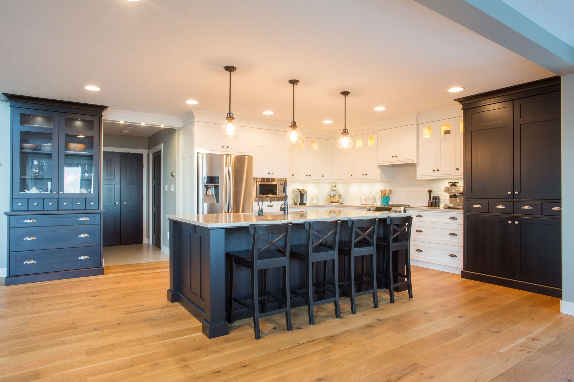A spacious kitchen with a dark blue island and stools, wood flooring, white cabinets, and matching dark pantry cabinets. A spacious kitchen with a dark blue island and stools, wood flooring, white cabinets, and matching dark pantry cabinets.