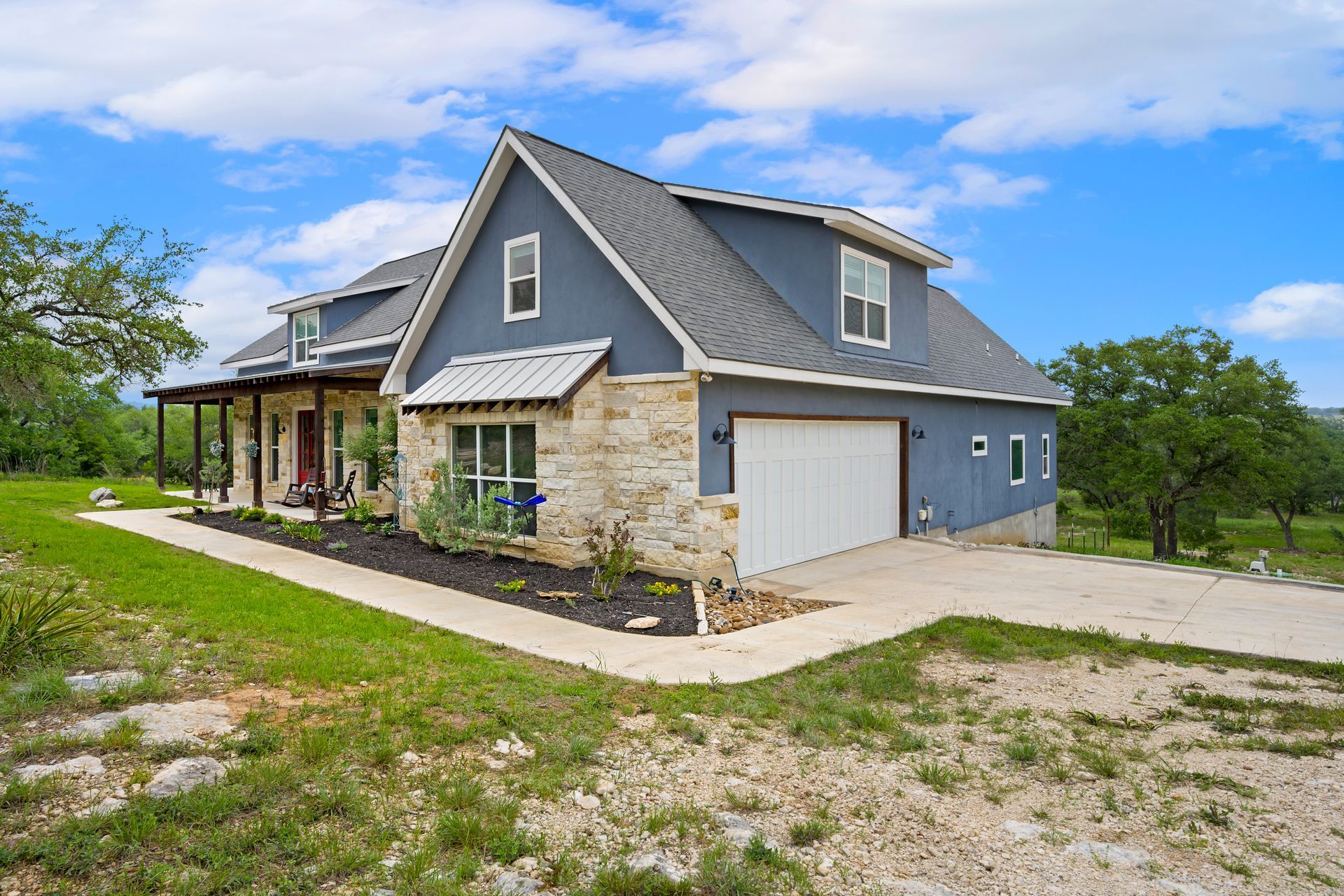 A two-story blue house with stone siding, a white garage, and a wrap-around porch set in a grassy landscape. A two-story blue house with stone siding, a white garage, and a wrap-around porch set in a grassy landscape.