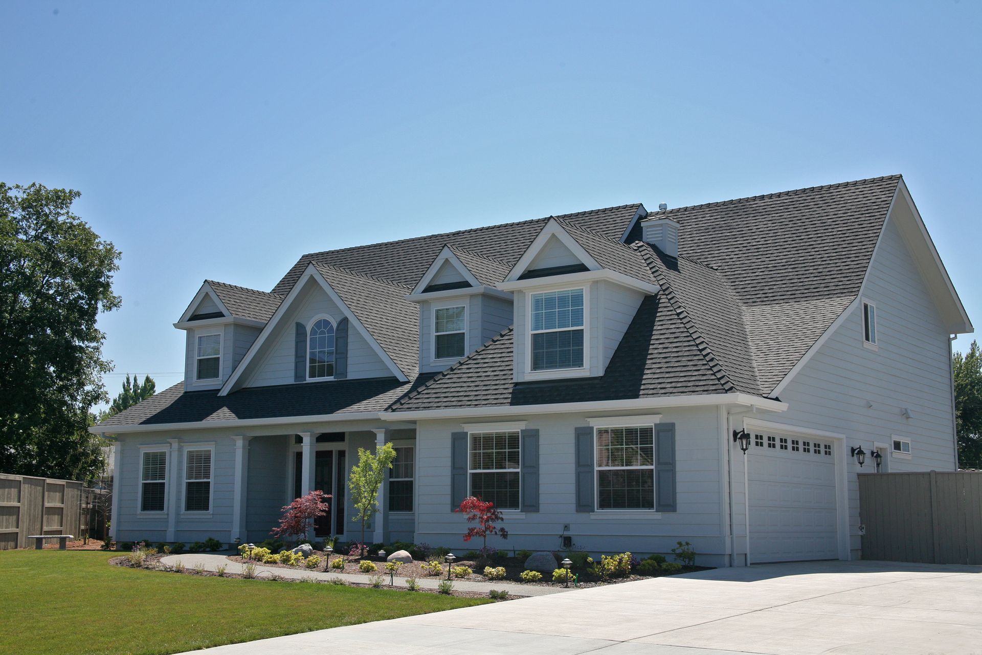 A light gray, two-story suburban house with a dark shingled roof, several dormer windows, and a paved driveway.