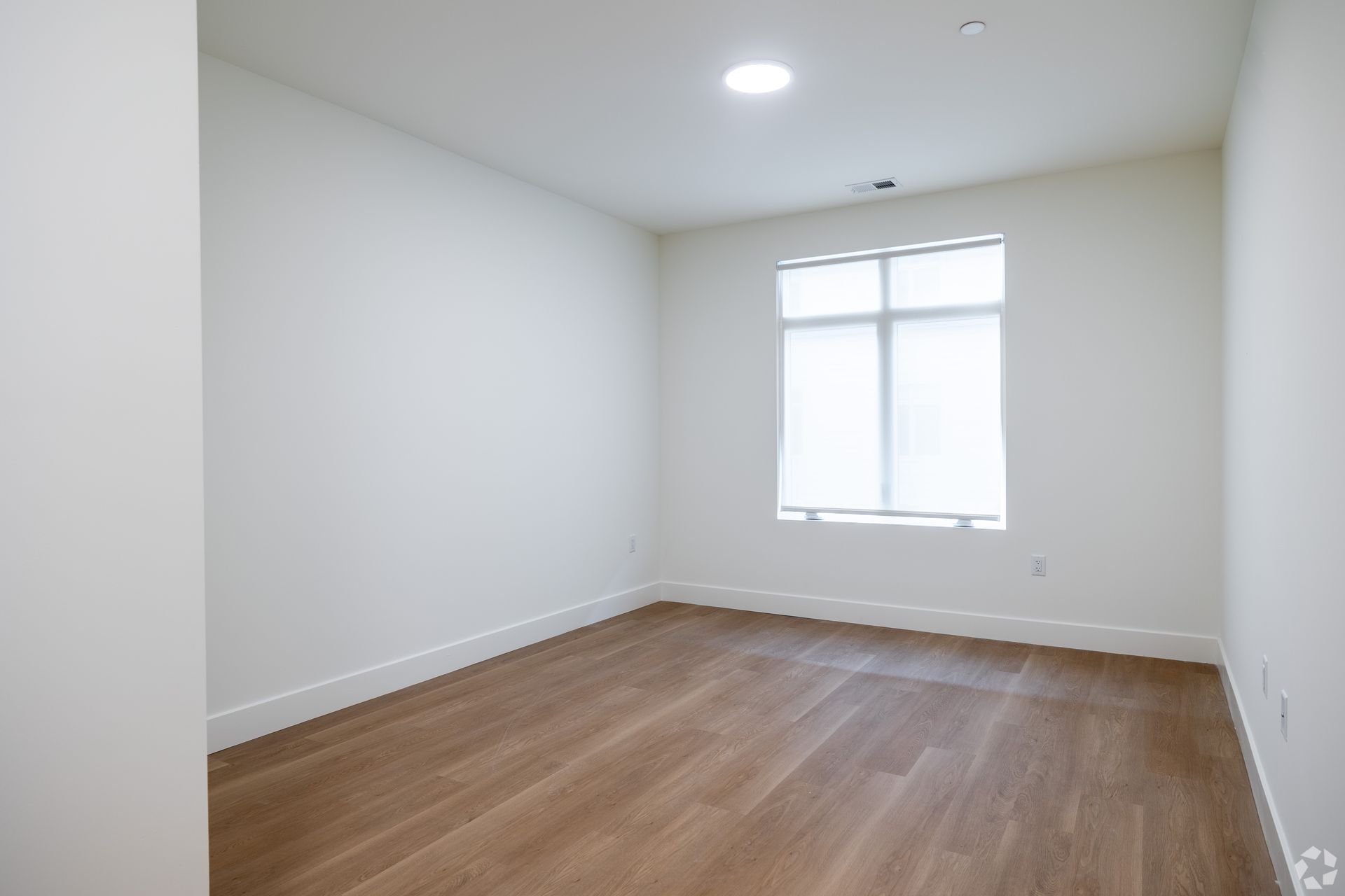 Empty room with light-colored wooden floor, white walls, and a window with a closed white shade.