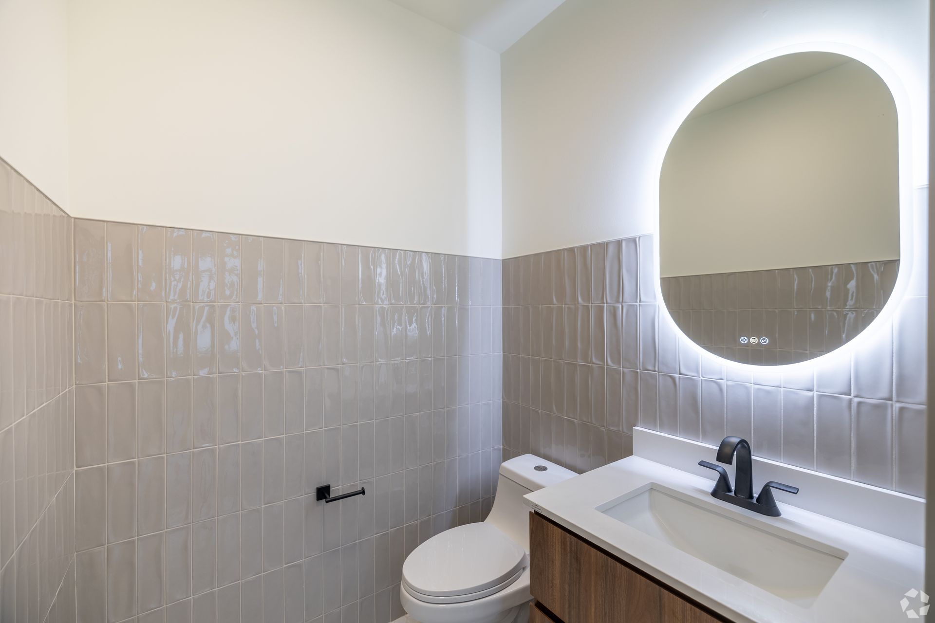 Modern bathroom with a light-up oval mirror, toilet, sink, and gray tiled accent wall.