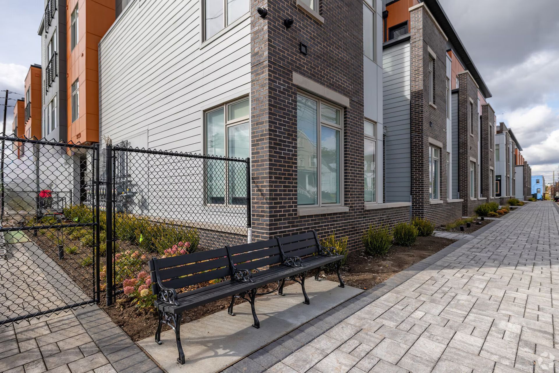 Black bench on a brick walkway in front of a modern building with silver siding and a black fence.
