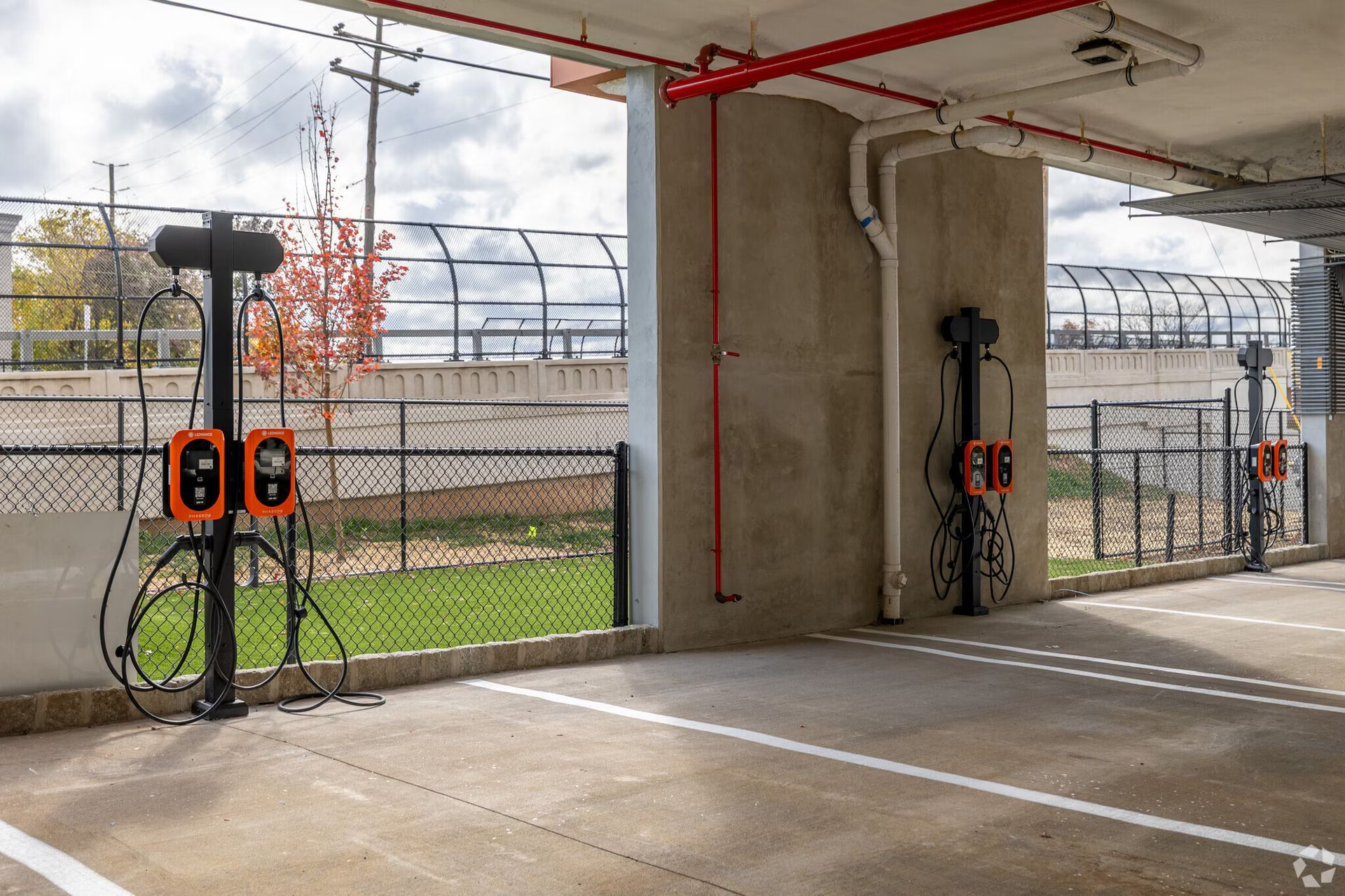 Electric vehicle charging stations in a parking garage. White concrete floor, red pipes overhead, black charging cables.