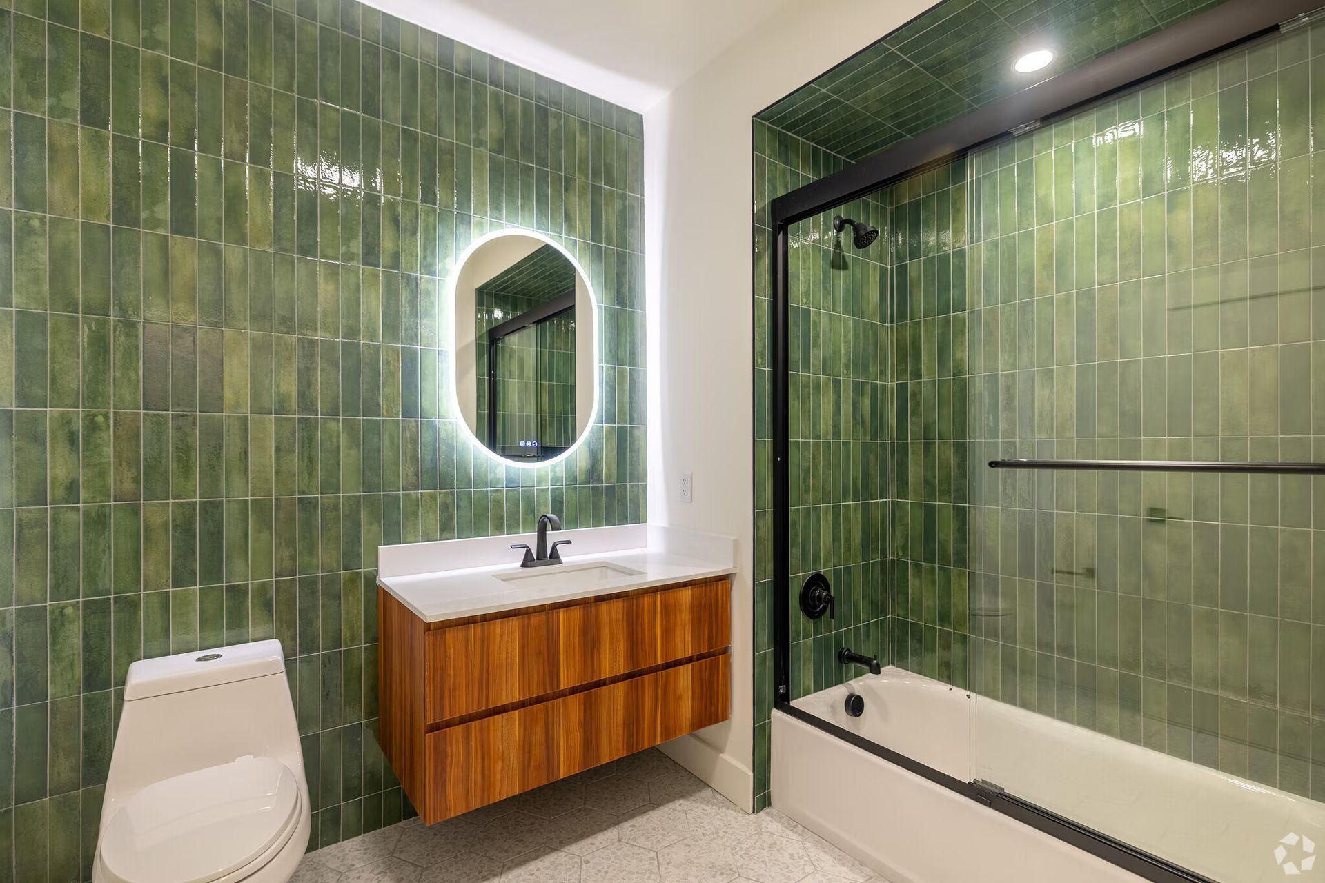 Green tiled bathroom with wooden vanity, oval mirror, and a bathtub.