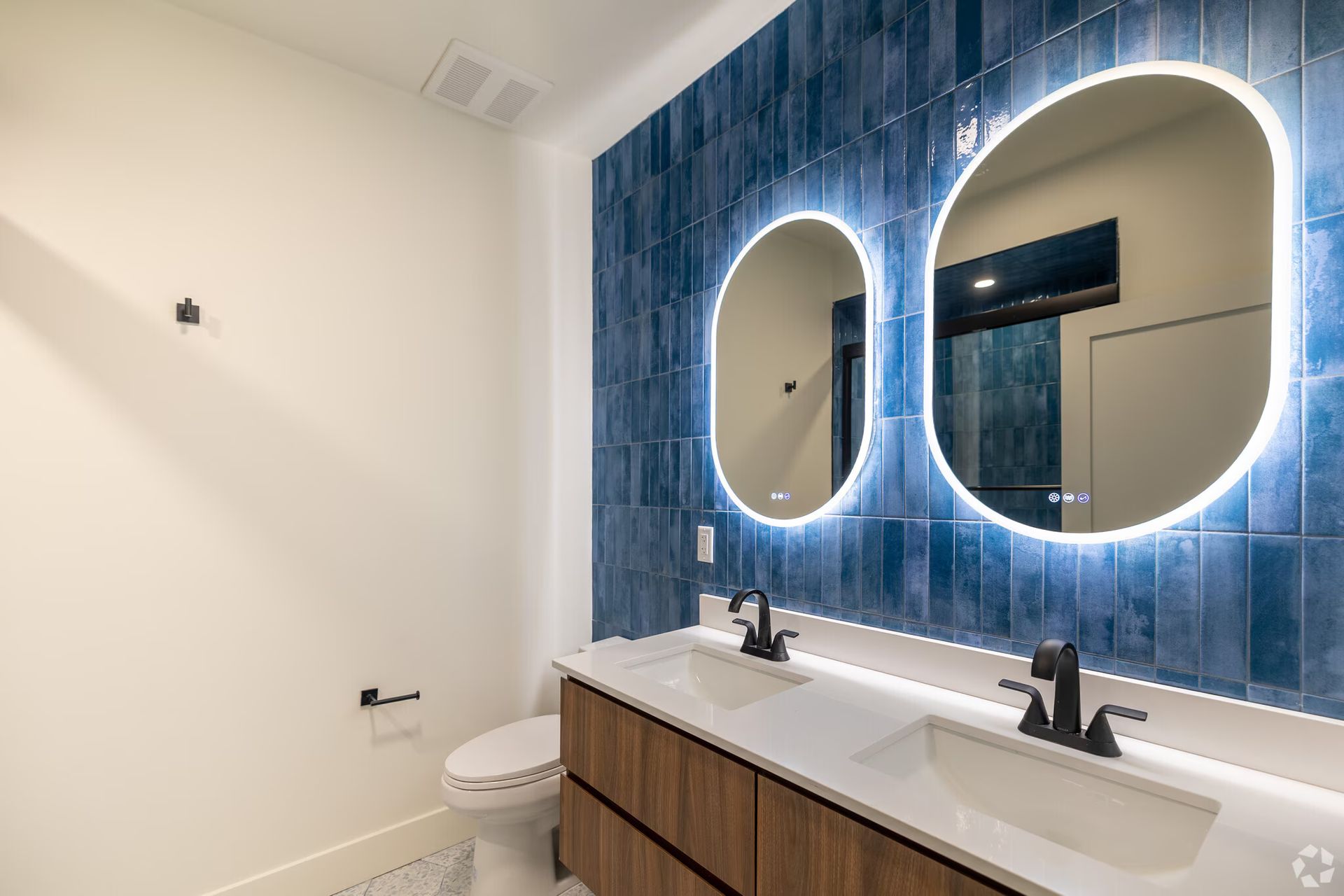 Bathroom with blue tiled accent wall, two oval mirrors with lights, double vanity, and toilet.