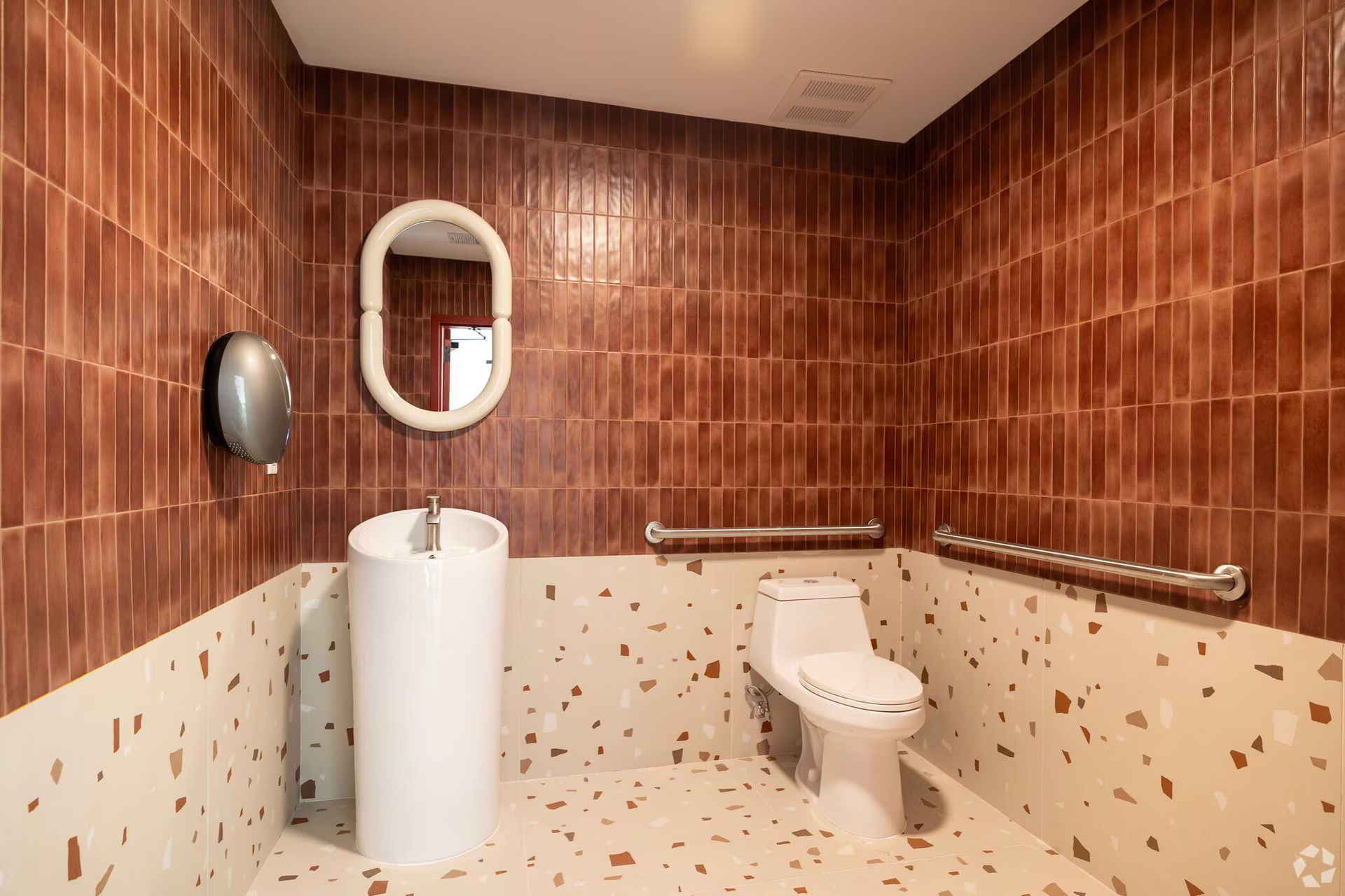 Brown-tiled bathroom with a white pedestal sink, toilet, and terrazzo flooring. Includes mirror, hand dryer, and grab bars.