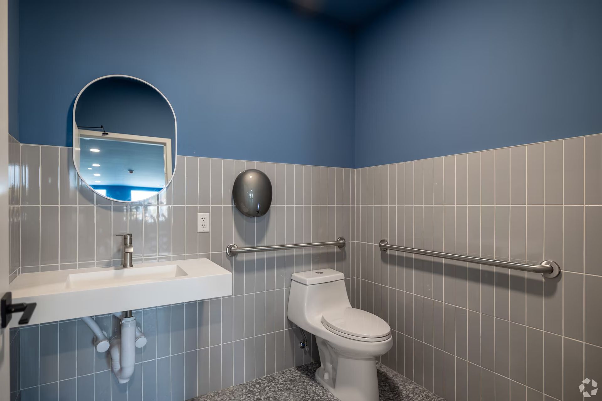 Accessible bathroom with a toilet, sink, mirror, and grab bars. Gray tiled walls and blue upper wall.