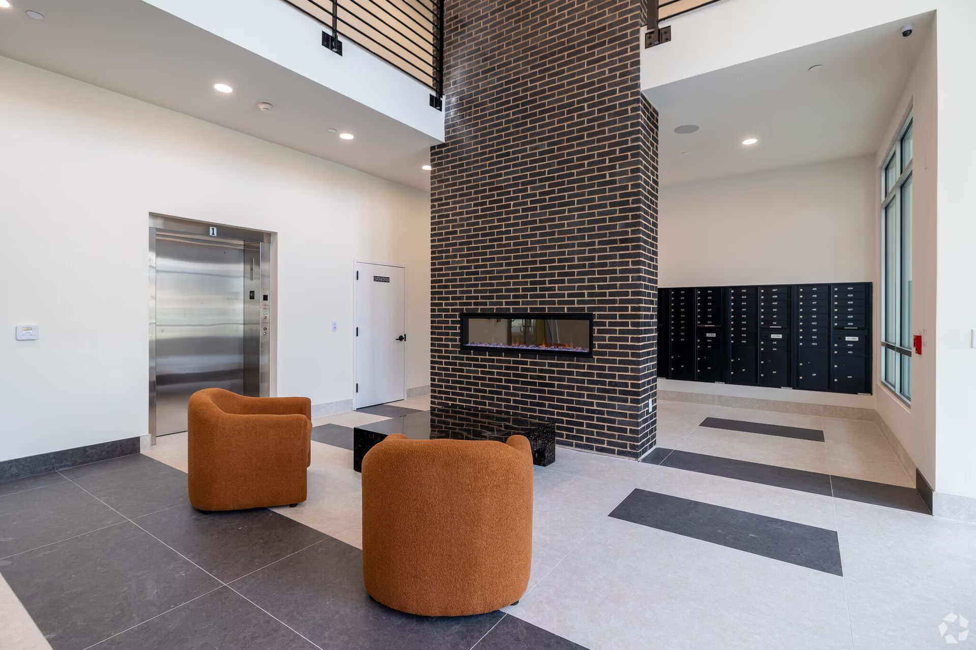 Lobby with orange armchairs, black brick fireplace, elevators, and black lockers.
