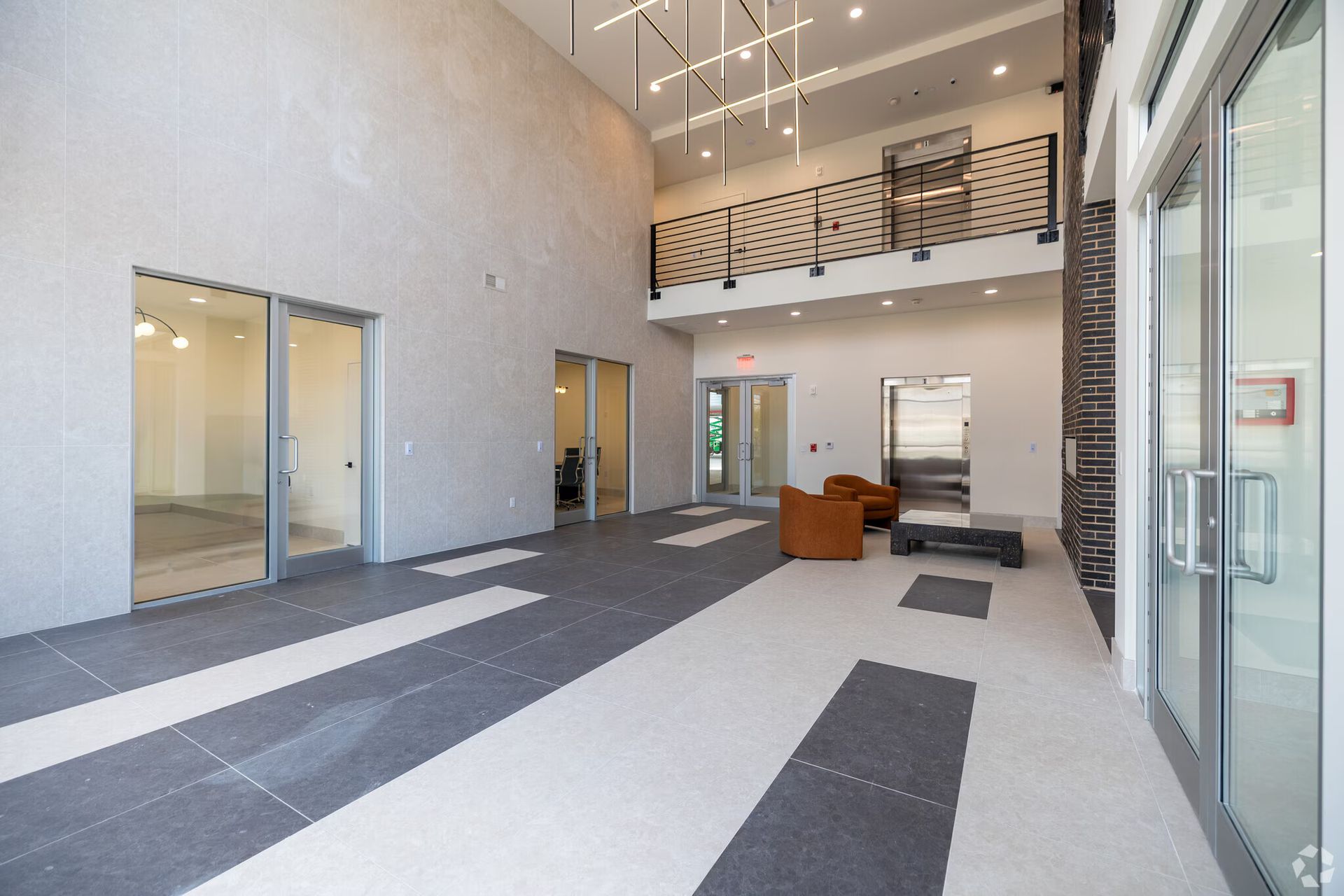 Modern building lobby with high ceilings, glass doors, seating area, and a second-floor balcony.