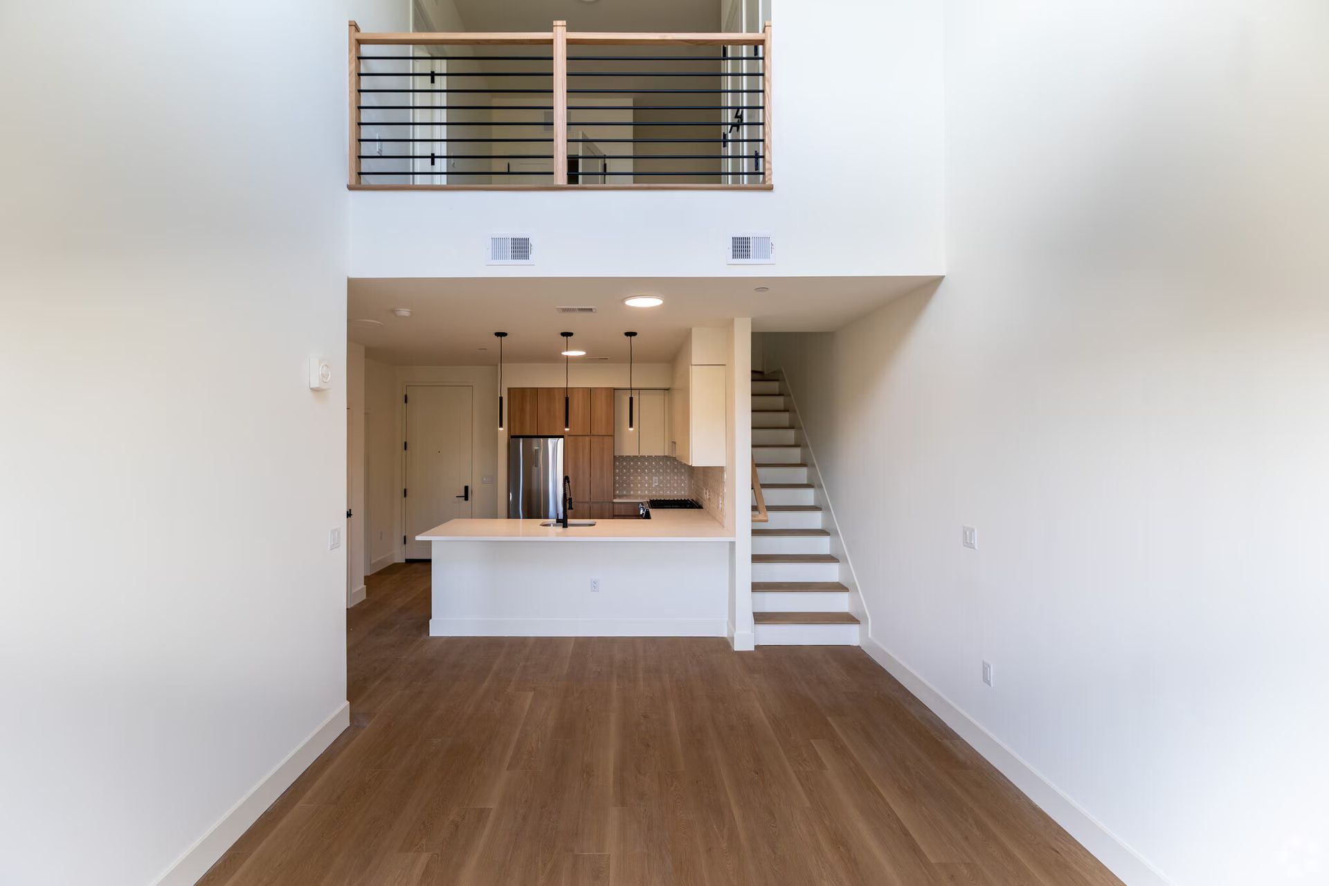 Interior view of a modern loft apartment with a kitchen, stairs, and a mezzanine level. Wooden floor and white walls.