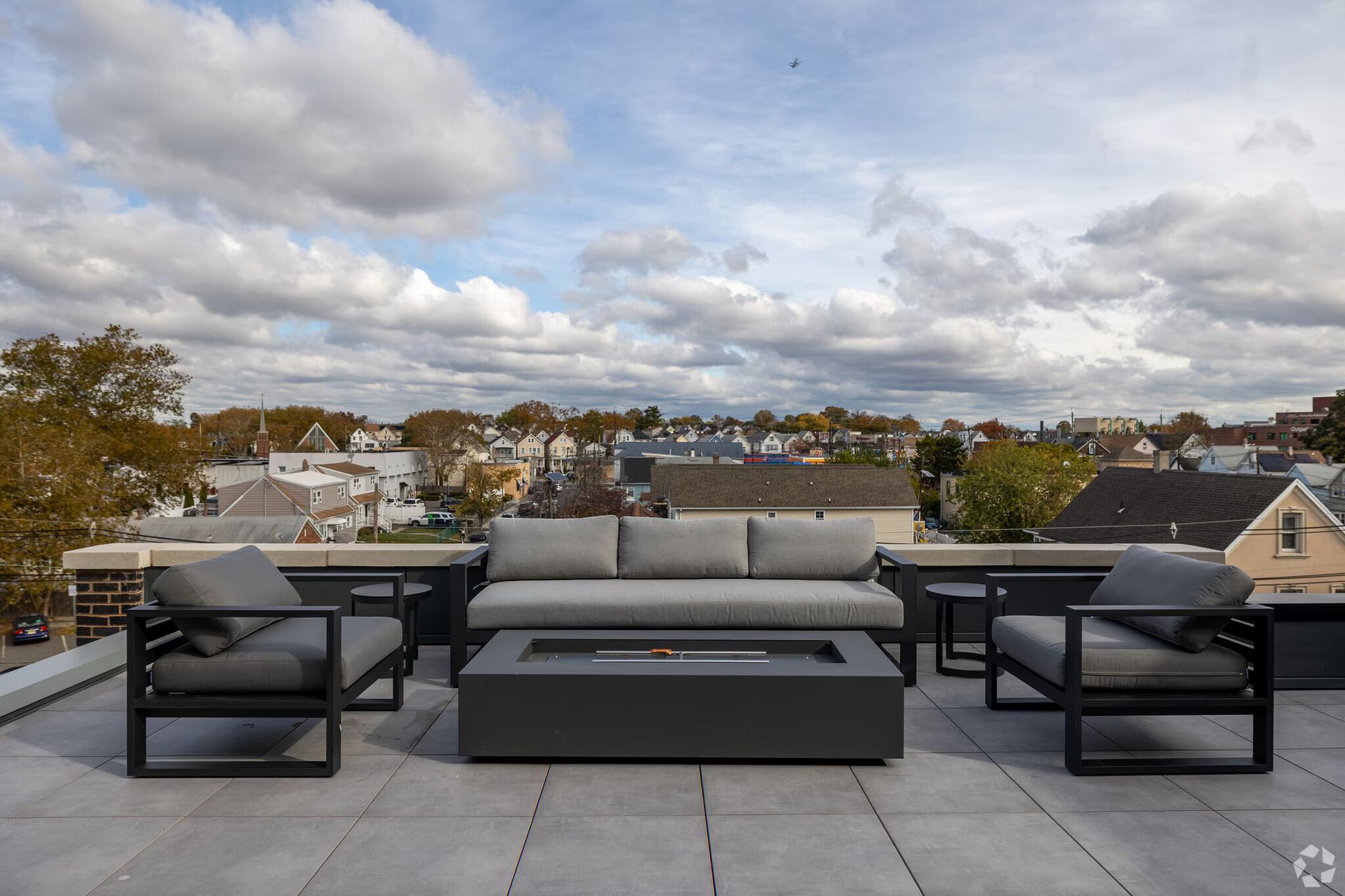 Rooftop patio with gray furniture, a fire pit, and a view of a neighborhood under a cloudy sky.