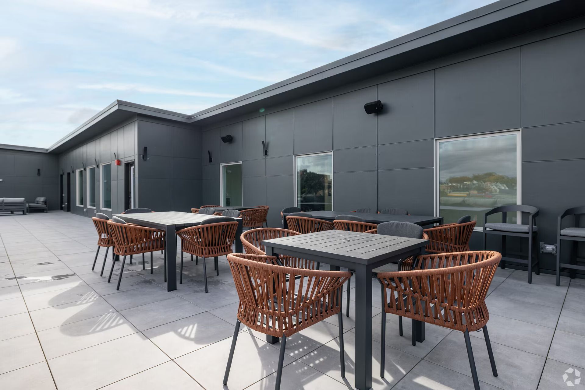 Outdoor patio with tables and chairs. Gray building exterior, cloudy sky.
