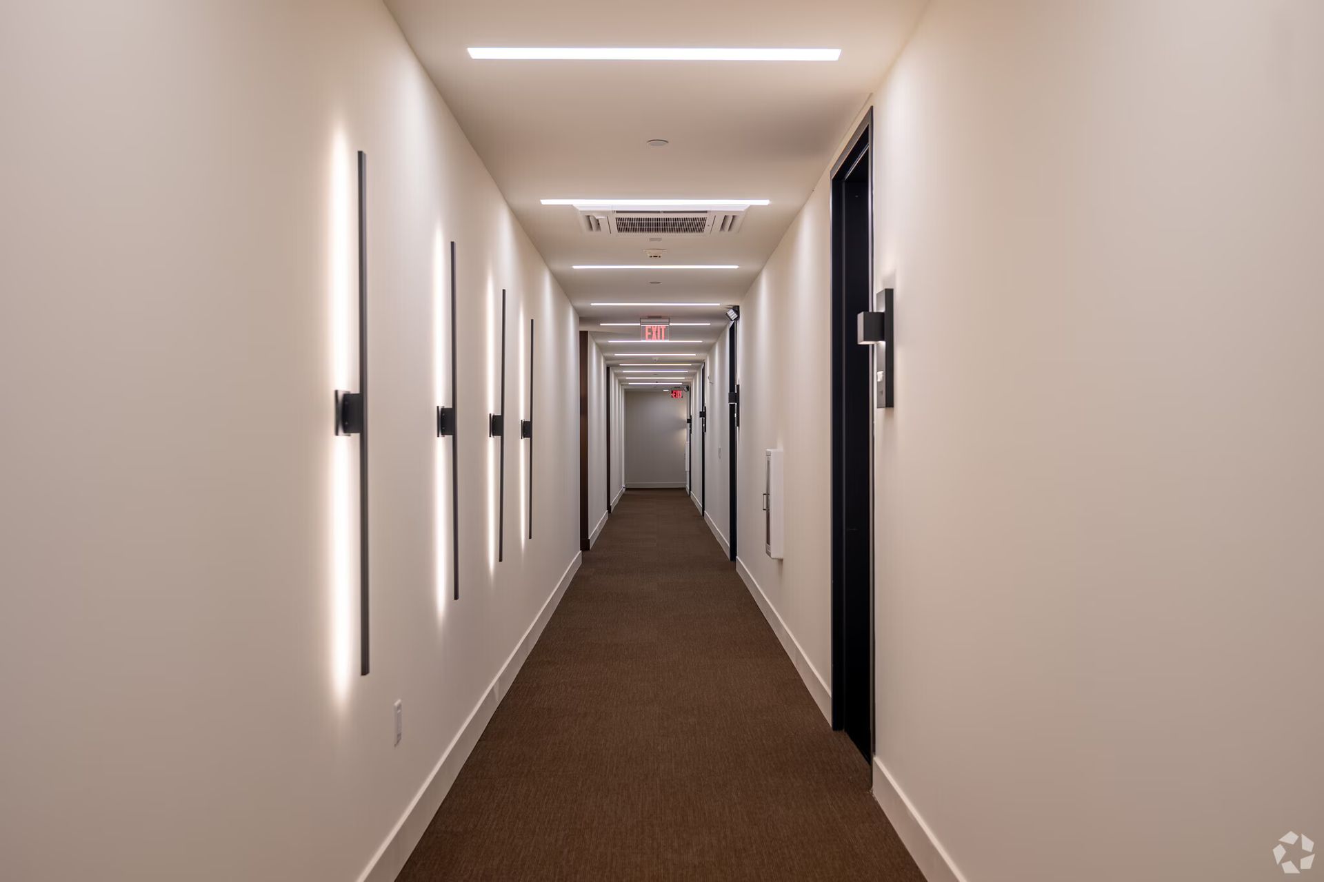 Long, narrow hallway with white walls, brown carpet, and modern lighting fixtures.