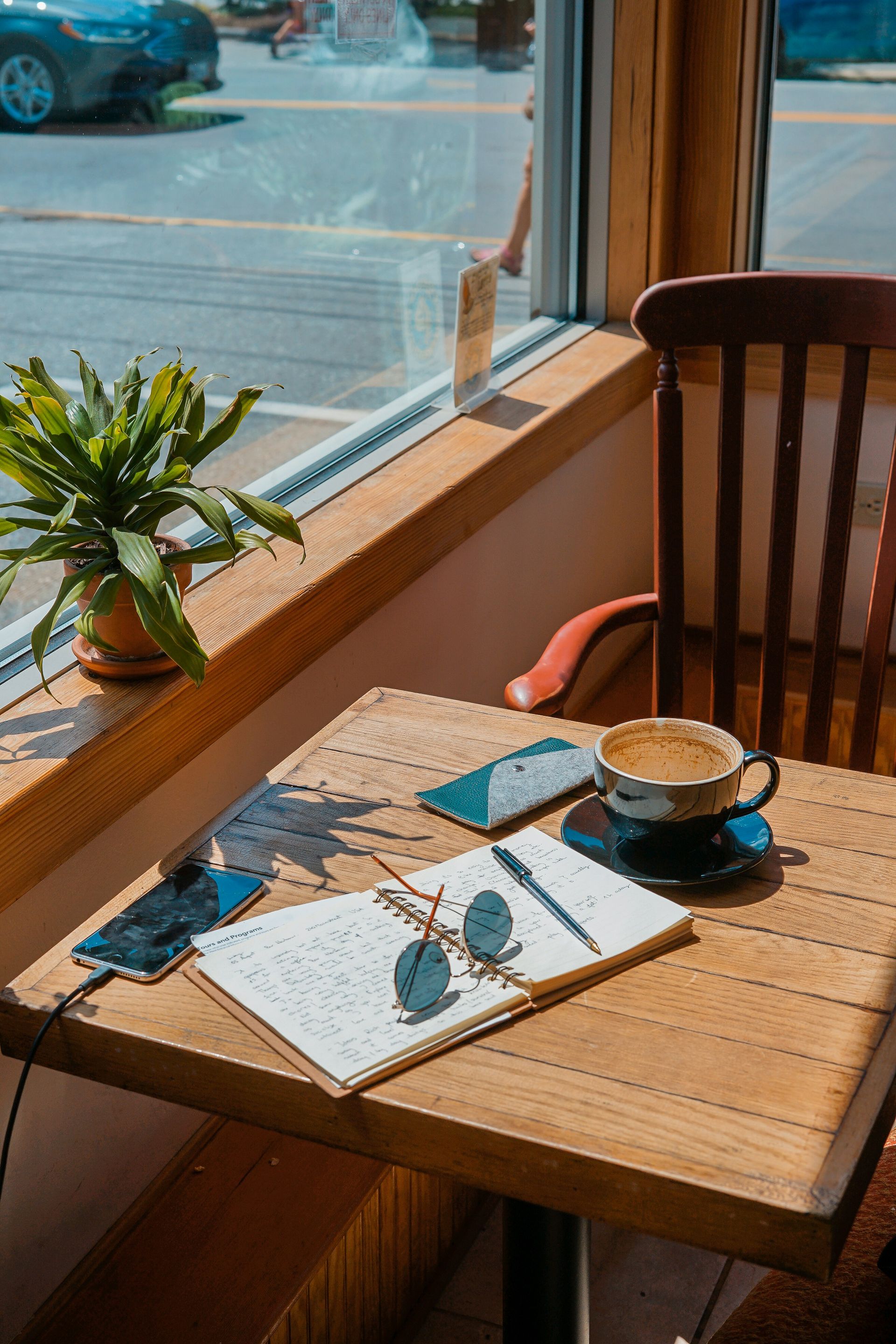 A wooden table with a cup of coffee , a notebook and sunglasses on it.