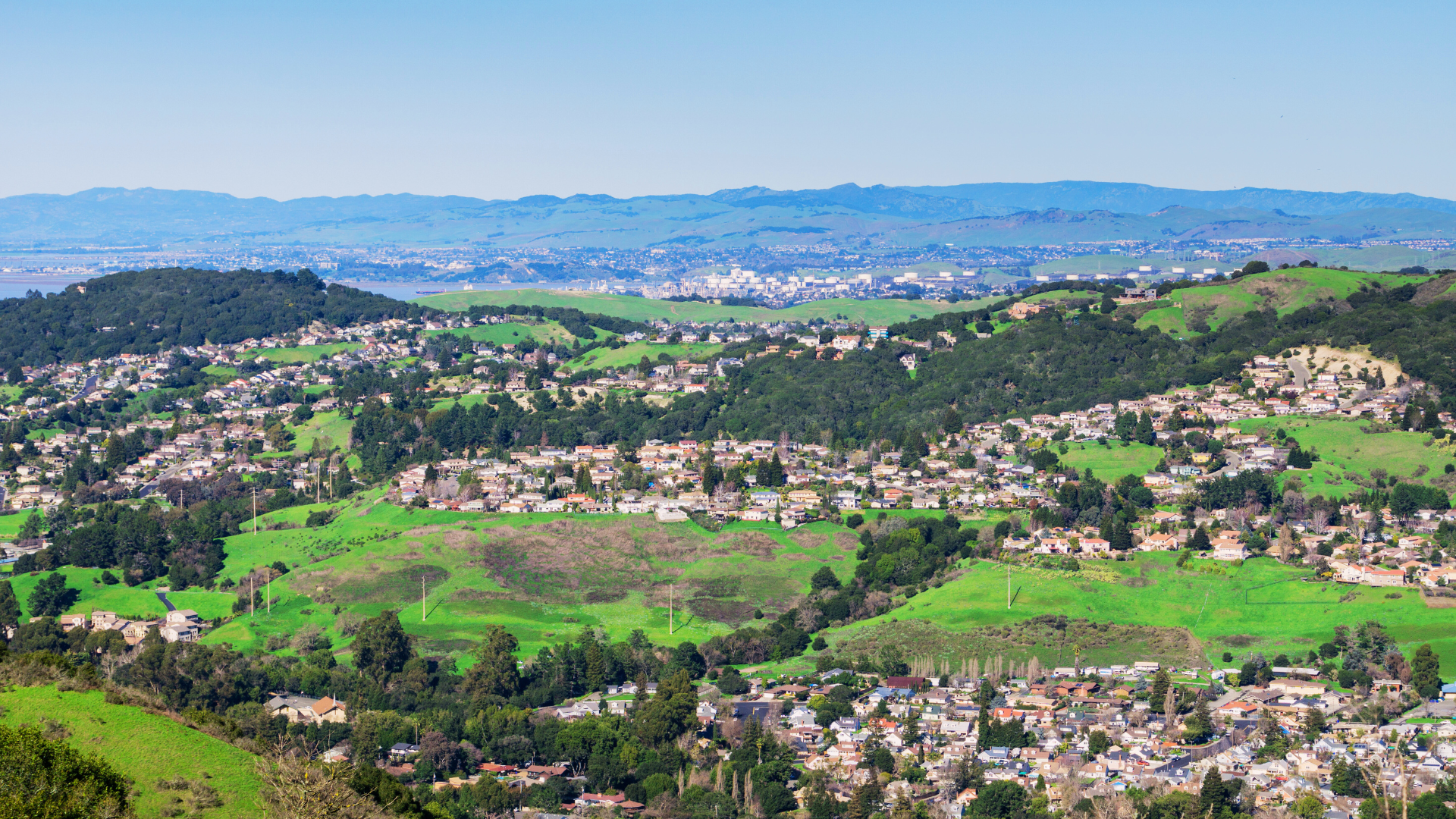 Panoramic view of a green hillside town with houses and distant mountains under a clear blue sky