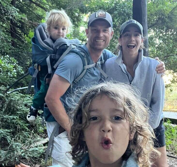 Family hiking on a wooded trail, with a child in front and three adults smiling behind them.