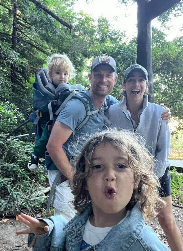 Family hiking selfie on a forest trail, with a child making a playful face in the foreground.