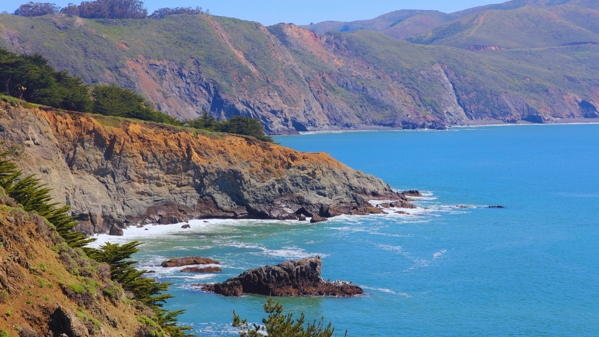 Rocky coastal cliffs above a bright blue ocean with a small cove and distant hills