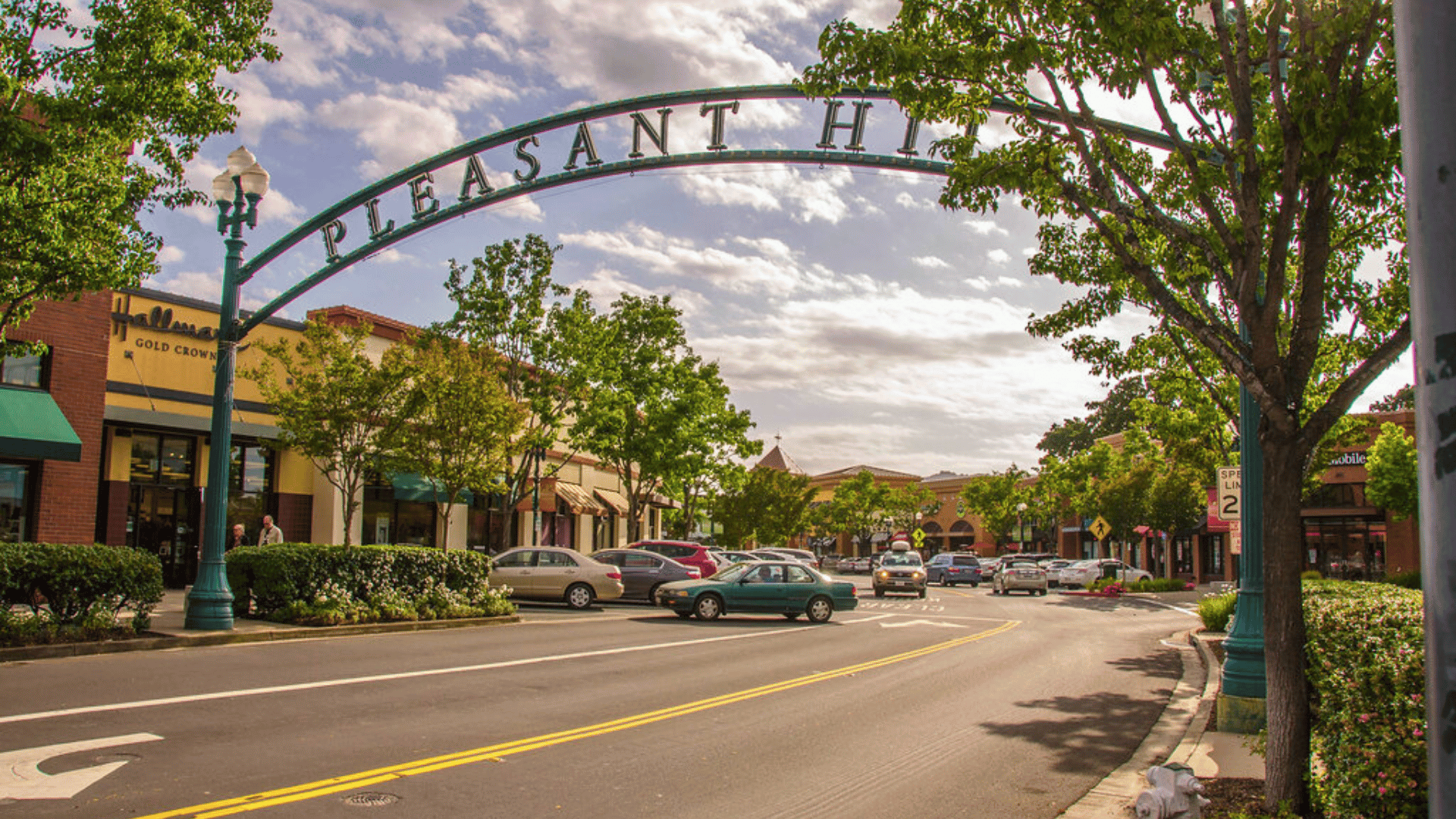 Arched “Pleasant Hill” sign over a tree-lined downtown street with storefronts and a few cars.