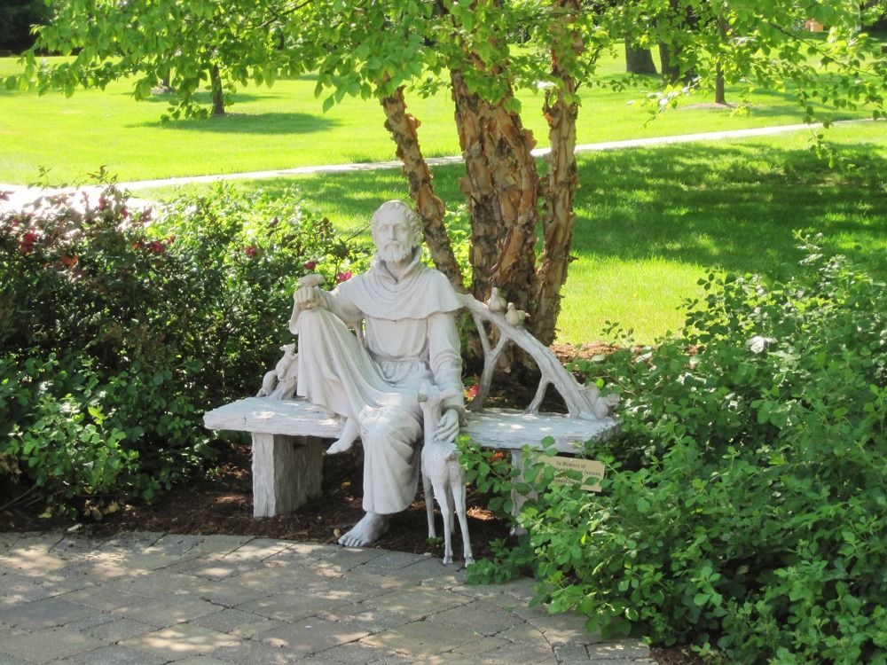 A white statue of a robed figure sits on a stone bench under a birch tree, surrounded by lush green shrubs in a park.