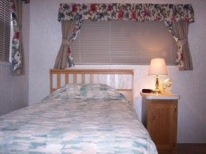 A twin bed with a patterned comforter next to a wooden nightstand with a lamp in a room with blinds and floral curtains.