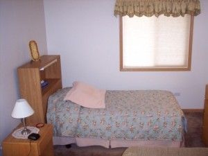 A single bed with a floral quilt and pink pillow, beside a wood nightstand with a lamp, under a window with valance.