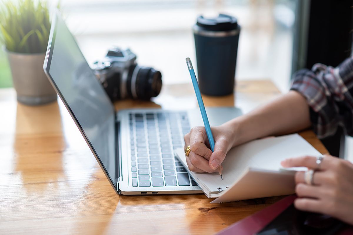 ou don't have to do this part alone Person writing notes in a notebook beside an open laptop with a camera and coffee on the desk