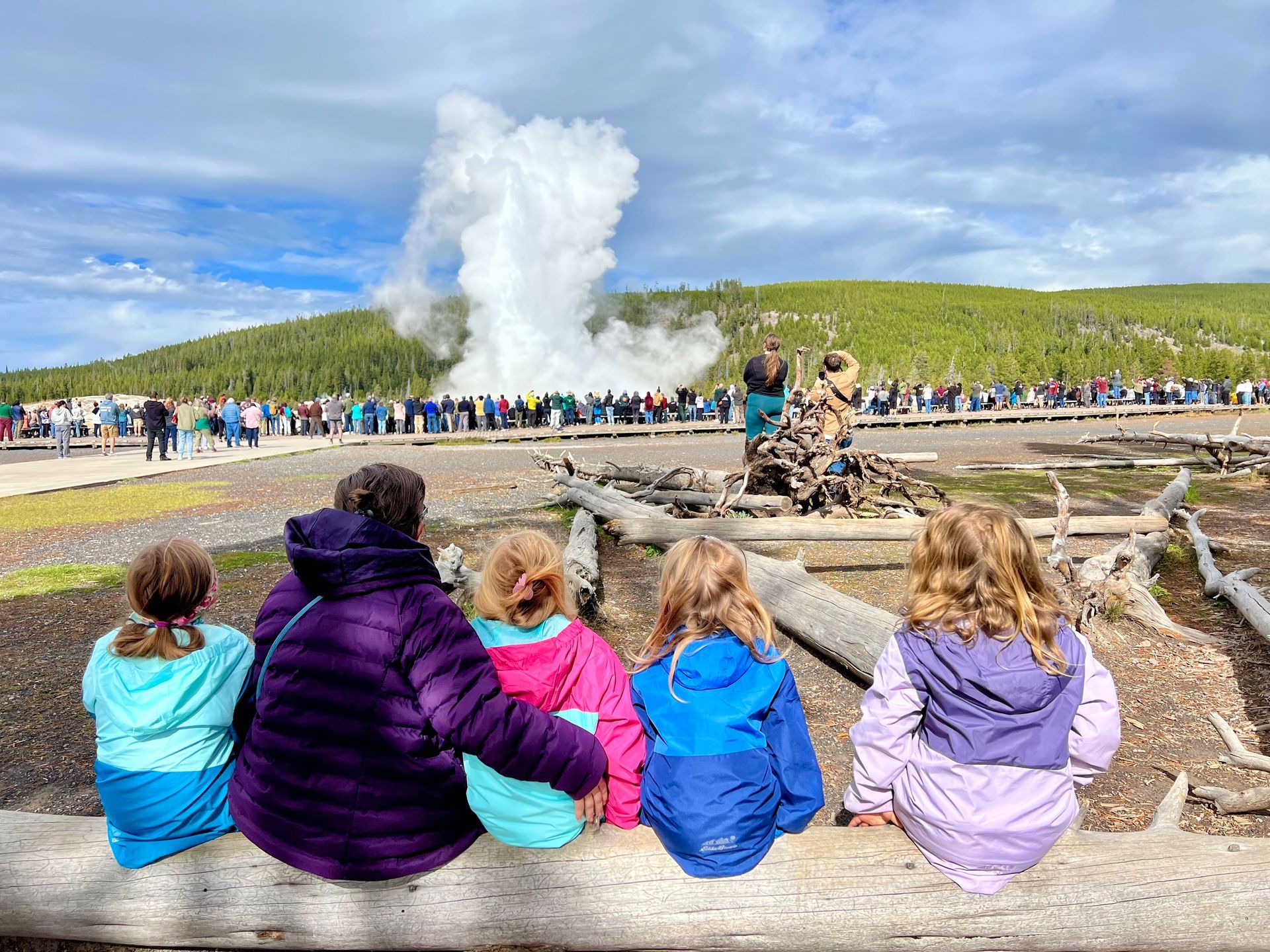 Family sitting on a log watching Old Faithful geyser erupt in Yellowstone National Park
