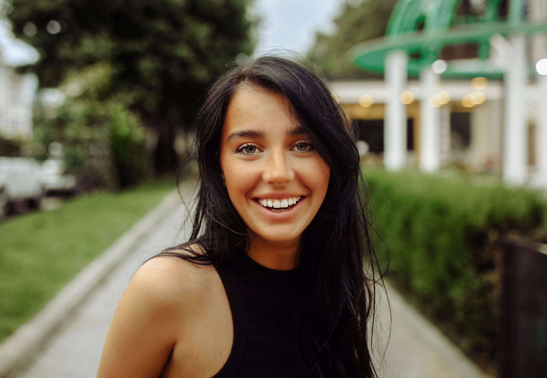 Smiling woman standing on a tree-lined path outdoors, with a blurred building in the background