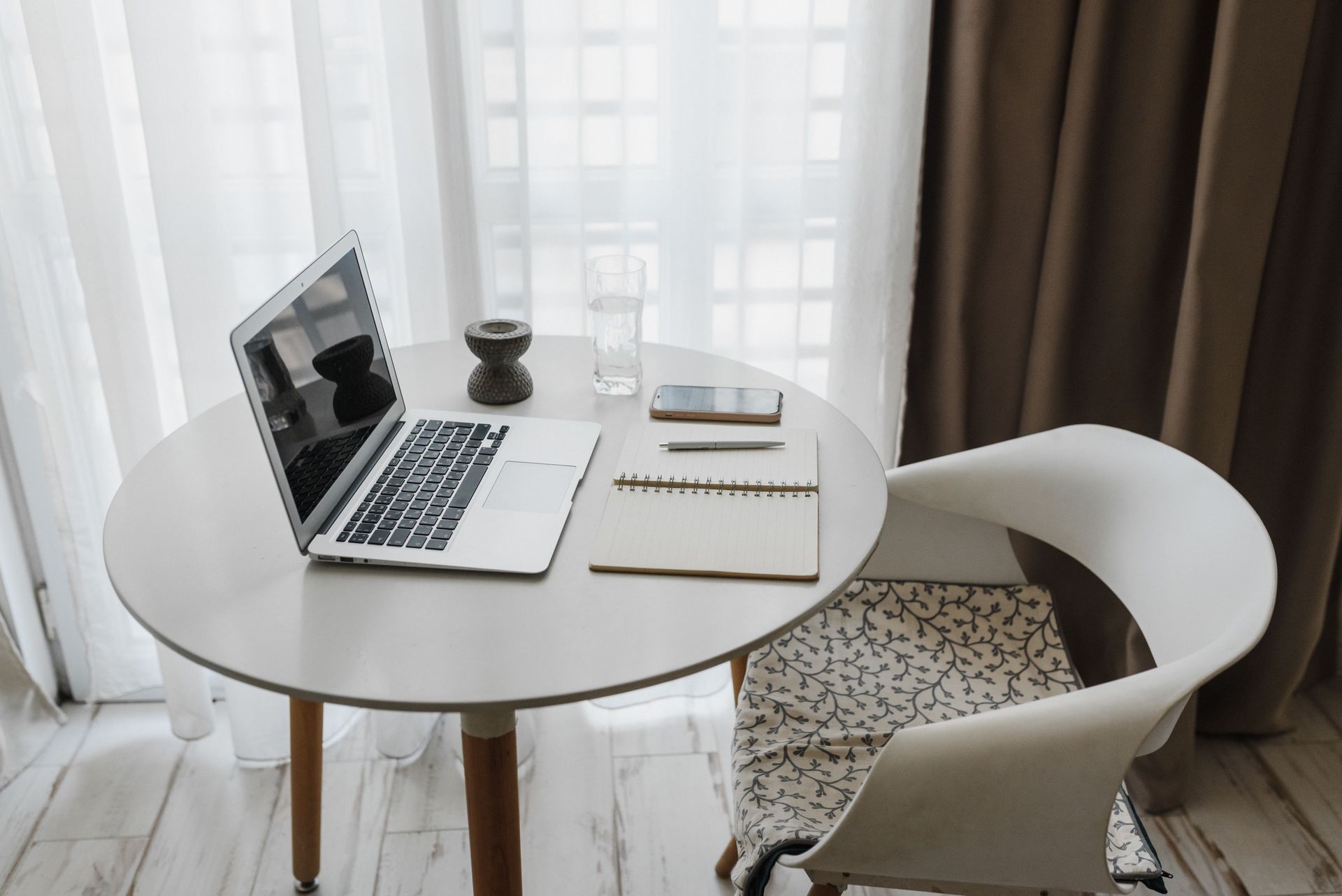 Minimal home office with a laptop, notebook, pen, and chair by a bright window