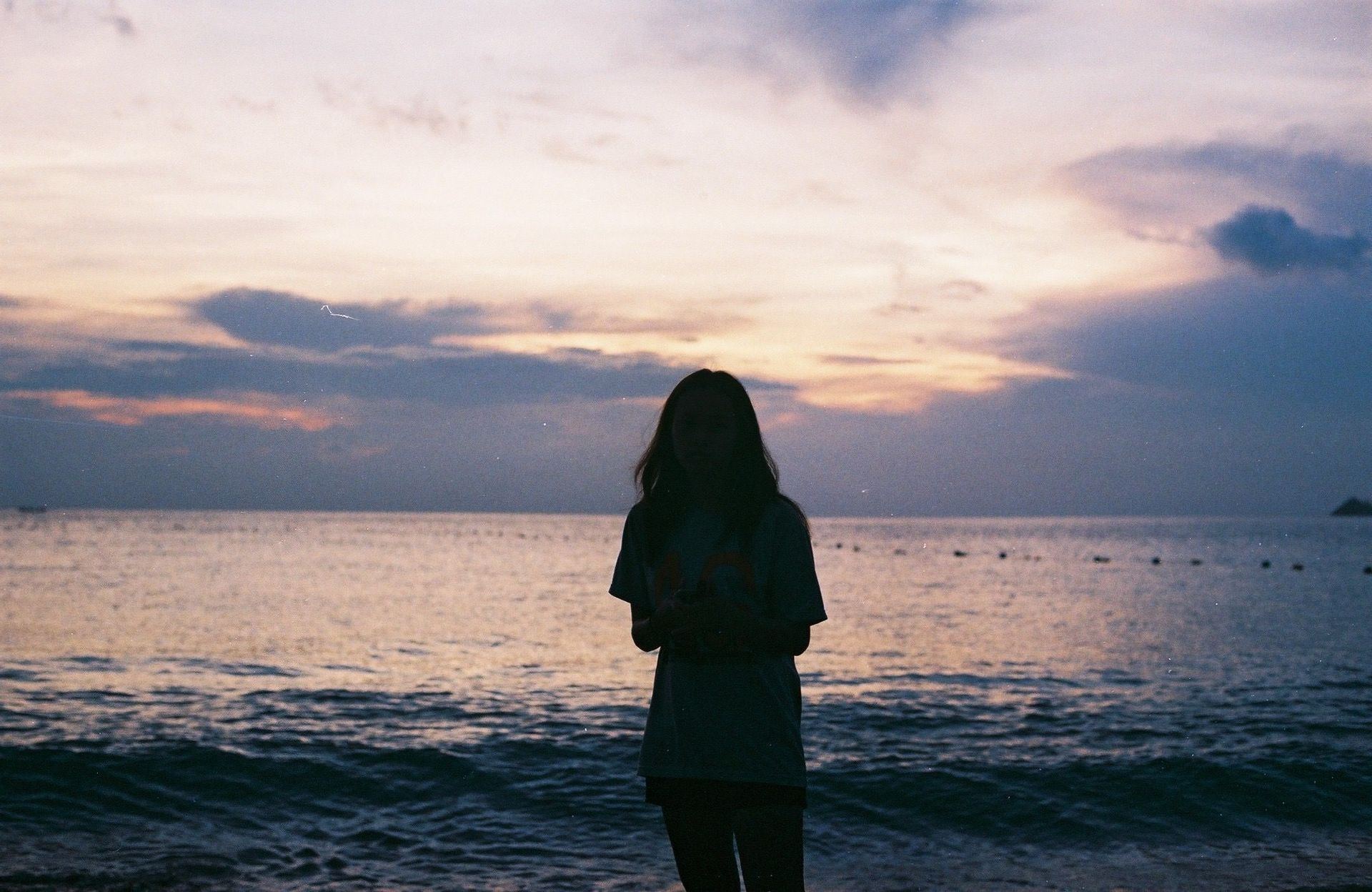 Silhouette of a person standing on a beach at sunset, facing the calm ocean and cloudy sky.