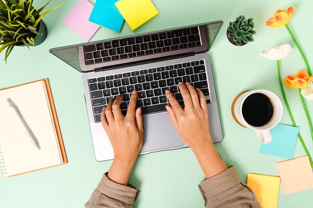 Calendar it and protect it Overhead flatlay of hands typing on a laptop surrounded by colorful sticky notes, a coffee cup, and plants on a green desk