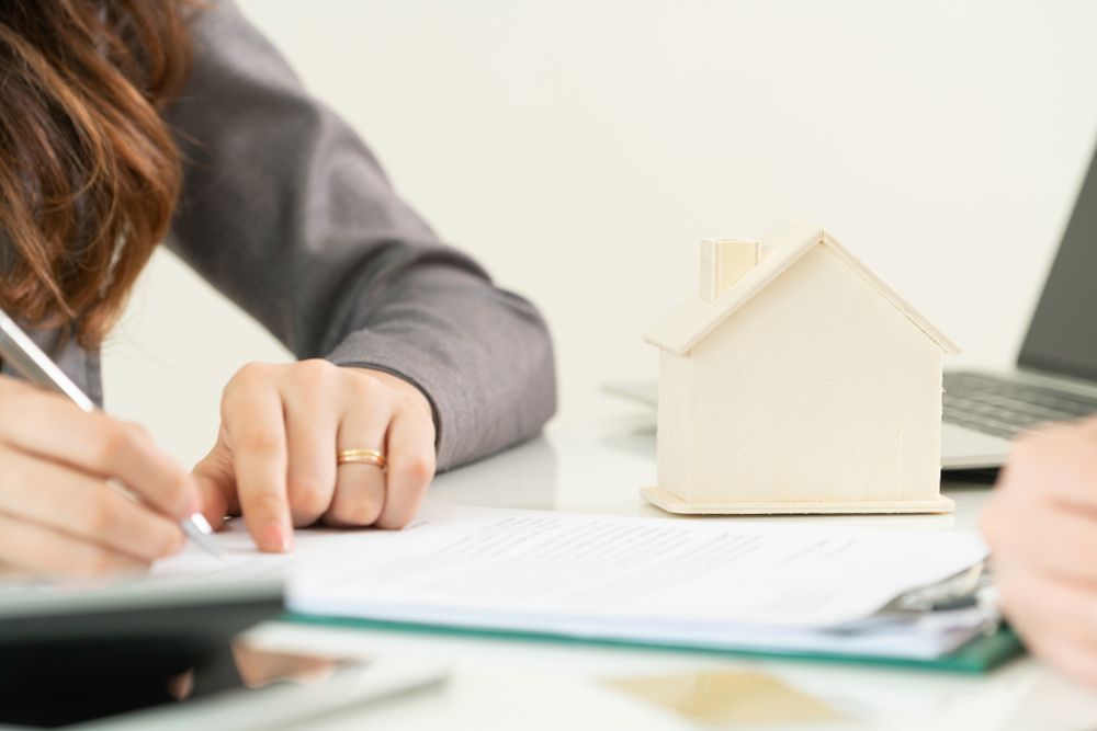A Woman Is Writing on A Piece of Paper Next to A Model House — A M Conveyancing Pty Ltd In Gold Coast, QLD