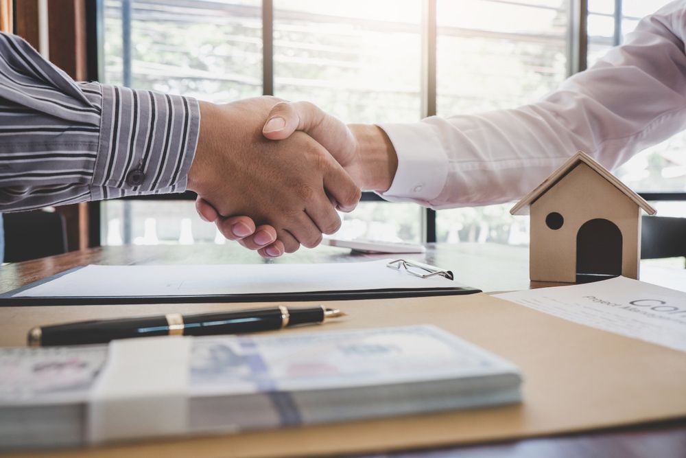 Two Men Are Shaking Hands Over a Table with A Small House in The Background — A M Conveyancing Pty Ltd In Gold Coast, QLD