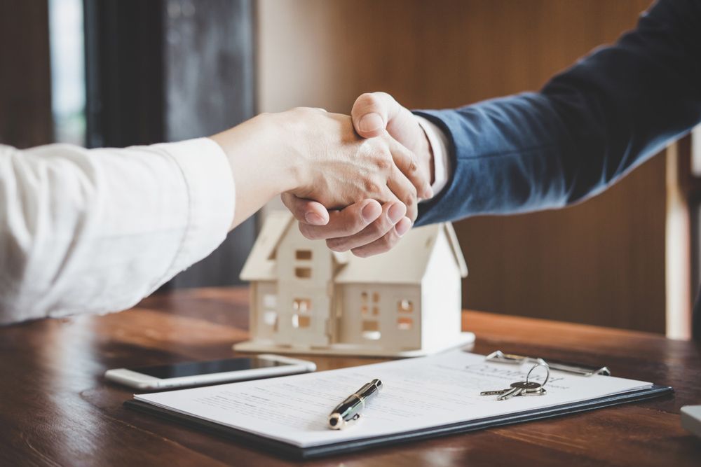 Two People Are Shaking Hands Over a Table with A Model House in The Background — A M Conveyancing Pty Ltd In Gold Coast, QLD