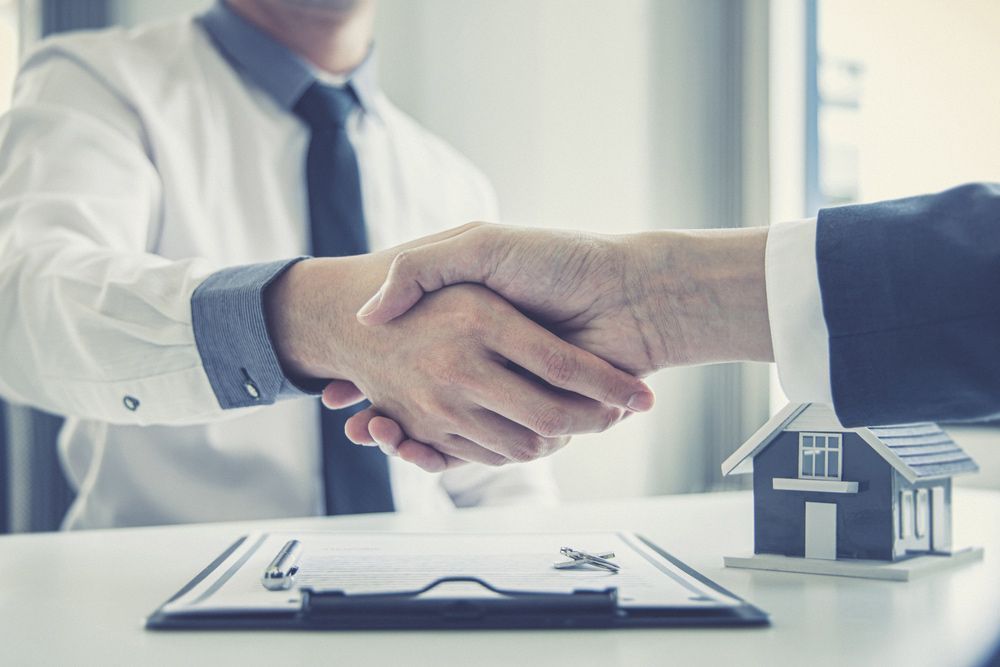 Two Men Are Shaking Hands Over a Table with A Model House in The Background — A M Conveyancing Pty Ltd In Tweed Heads, NSW