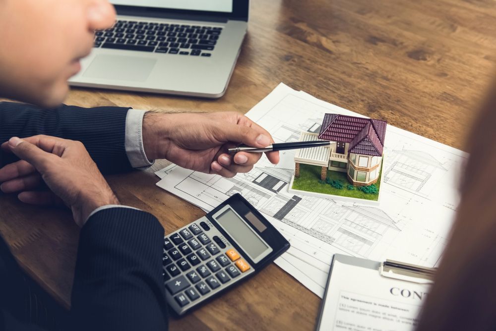A Man Is Sitting at A Table with A Calculator and A Model House — A M Conveyancing Pty Ltd In Palm Beach, QLD
