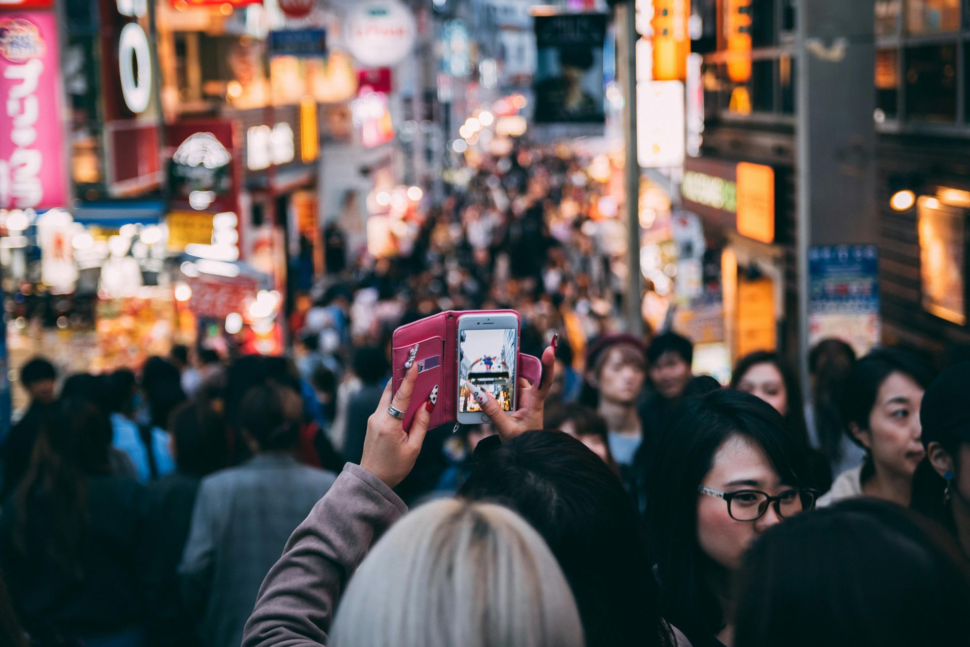 Crowd in a brightly lit city street, person taking photo with two phones.