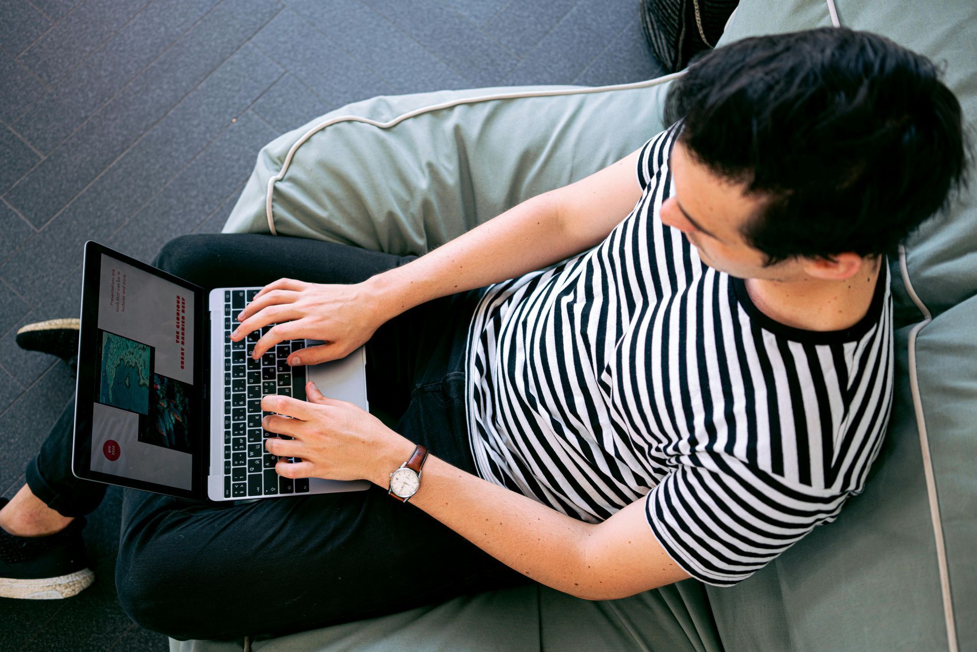 Person typing on laptop while sitting on a beanbag chair, wearing striped shirt and watch.
