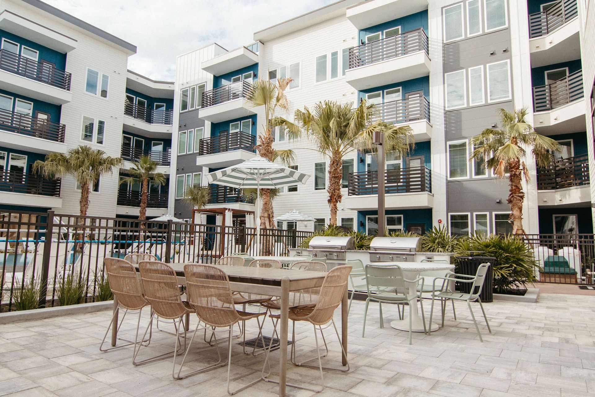 Outdoor patio area with tables, chairs, grills, and a pool in a multi-story apartment complex with palm trees.