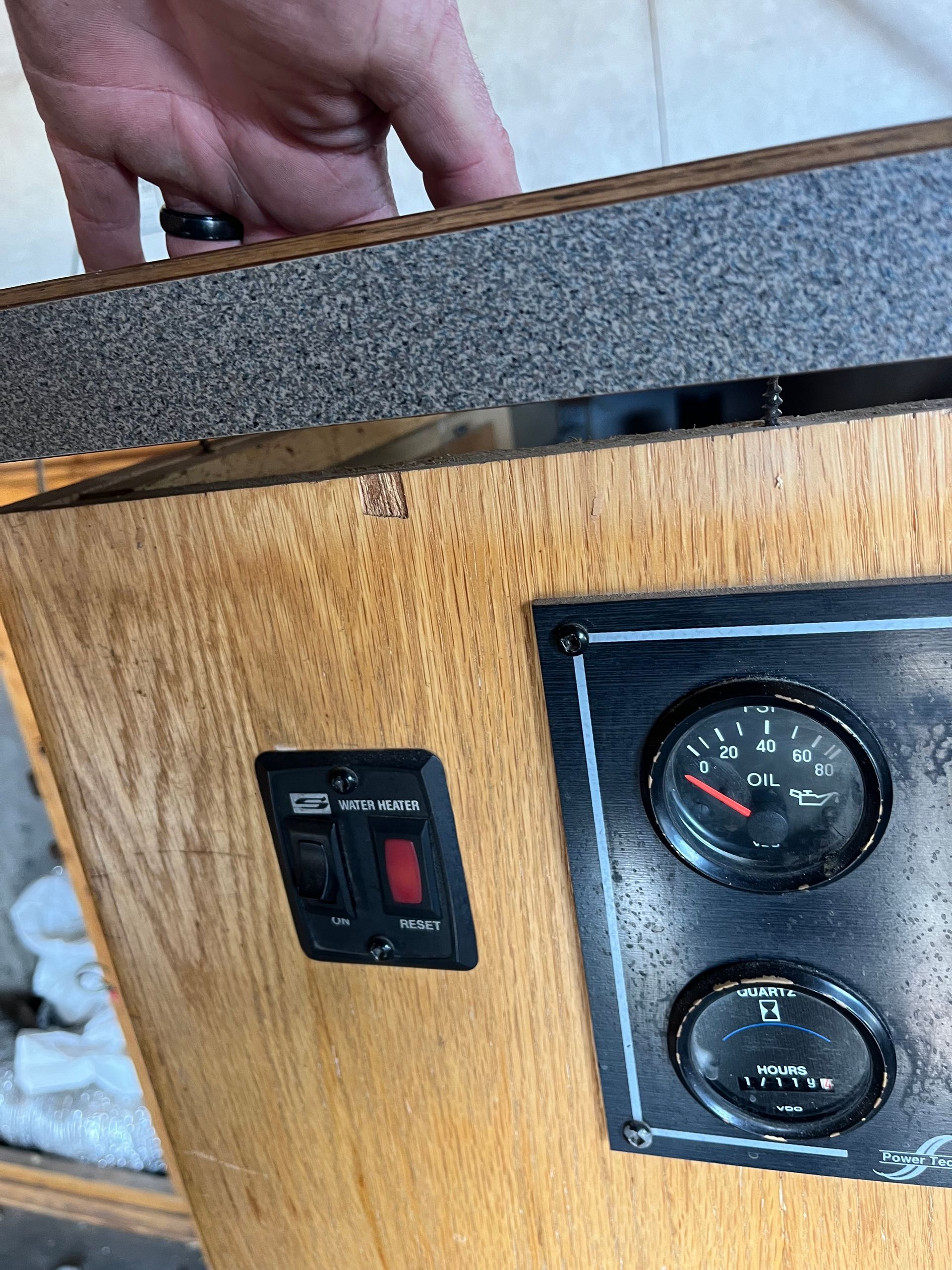 Hand above wood cabinet with gauges and a switch. Black gauges show markings, a red switch is on.
