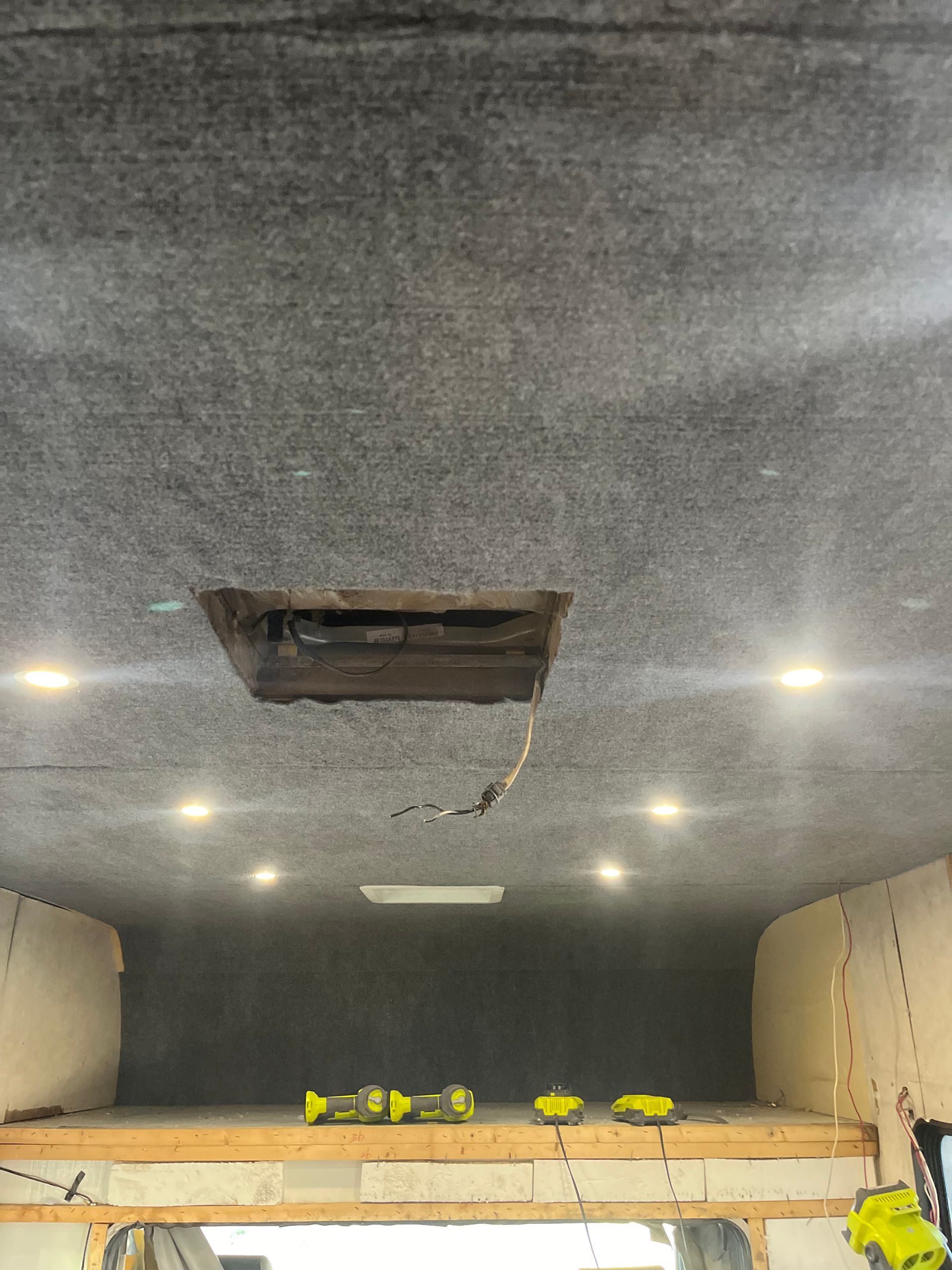 Interior view of a vehicle ceiling, with a square opening, lights, and tools visible below a bed platform.
