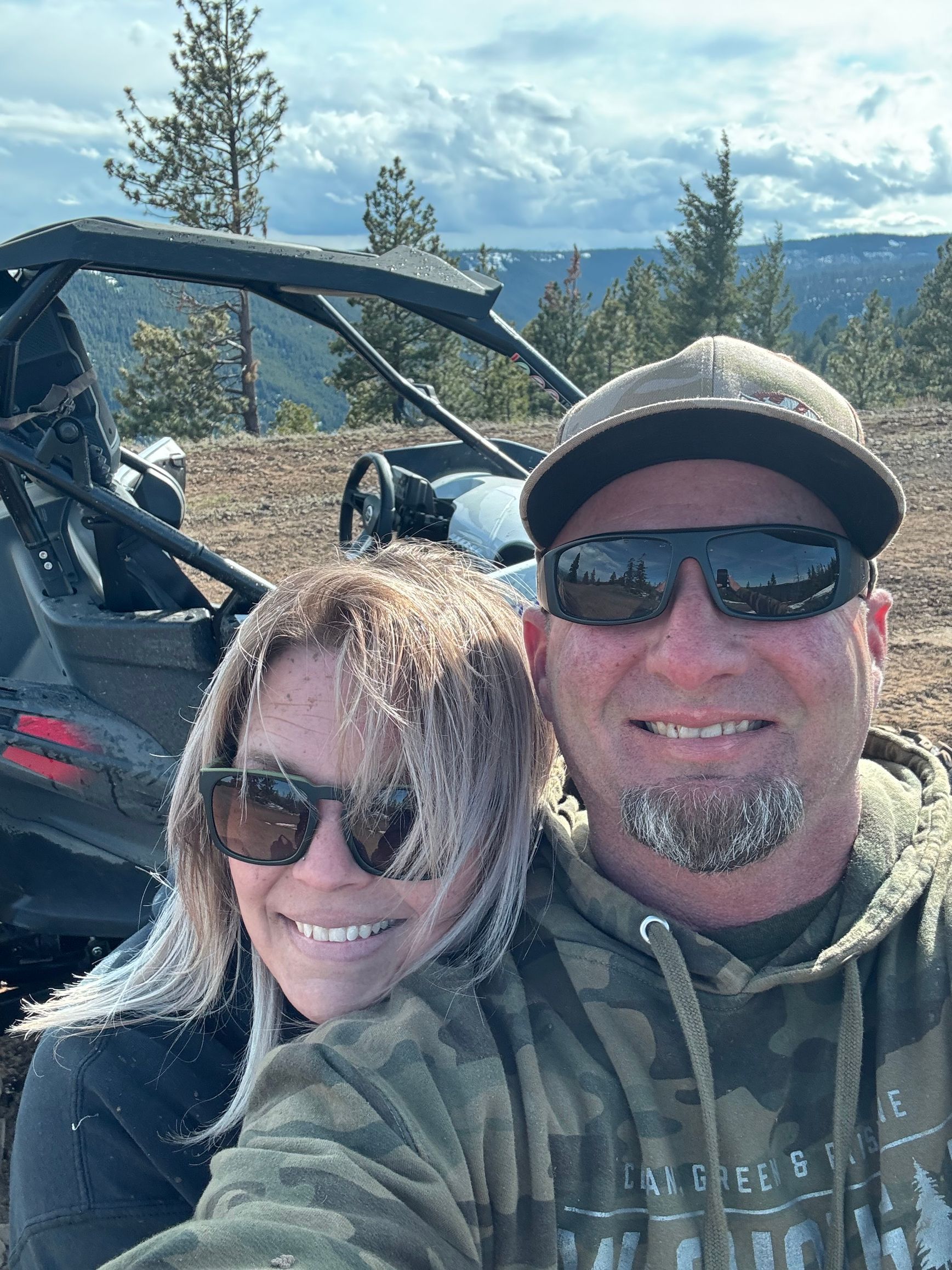 Couple wearing sunglasses smile for selfie with an off-road vehicle and scenic background.