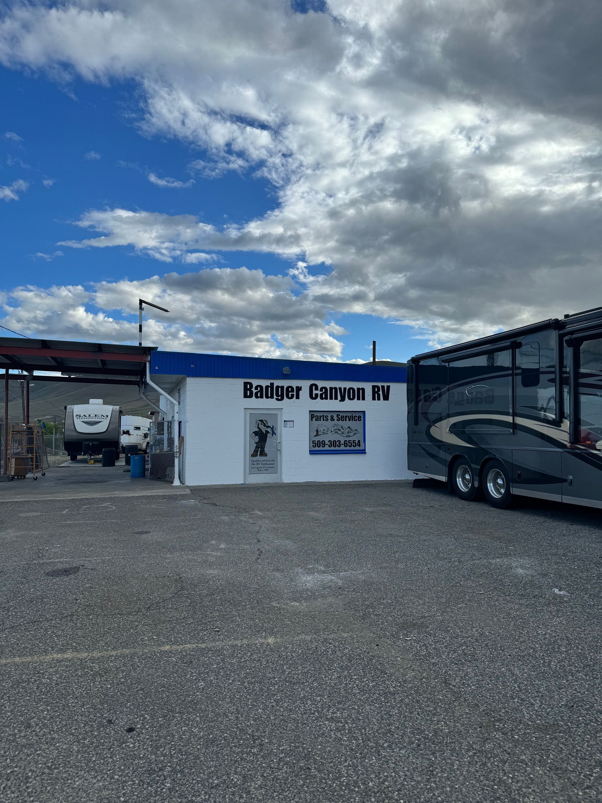 A small, white RV repair shop under a cloudy sky. A motorhome and RVs sit outside.