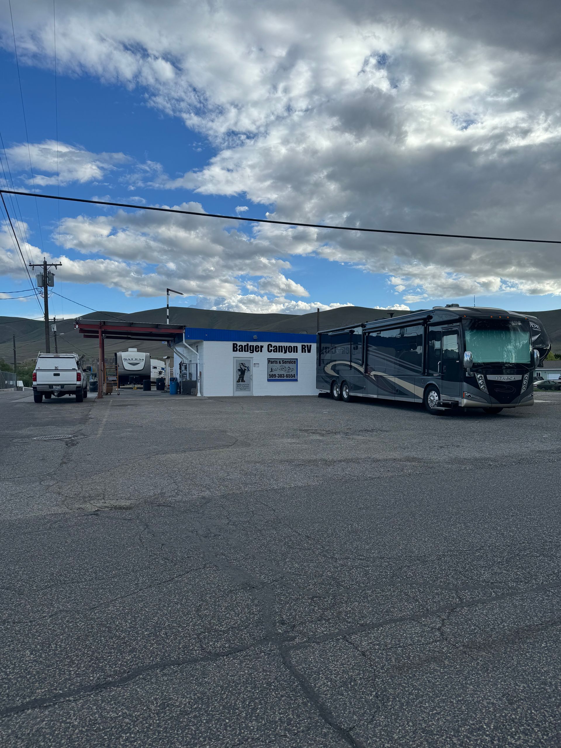 A small white building with a blue trim sits in a parking lot. An RV and several vehicles are parked nearby.