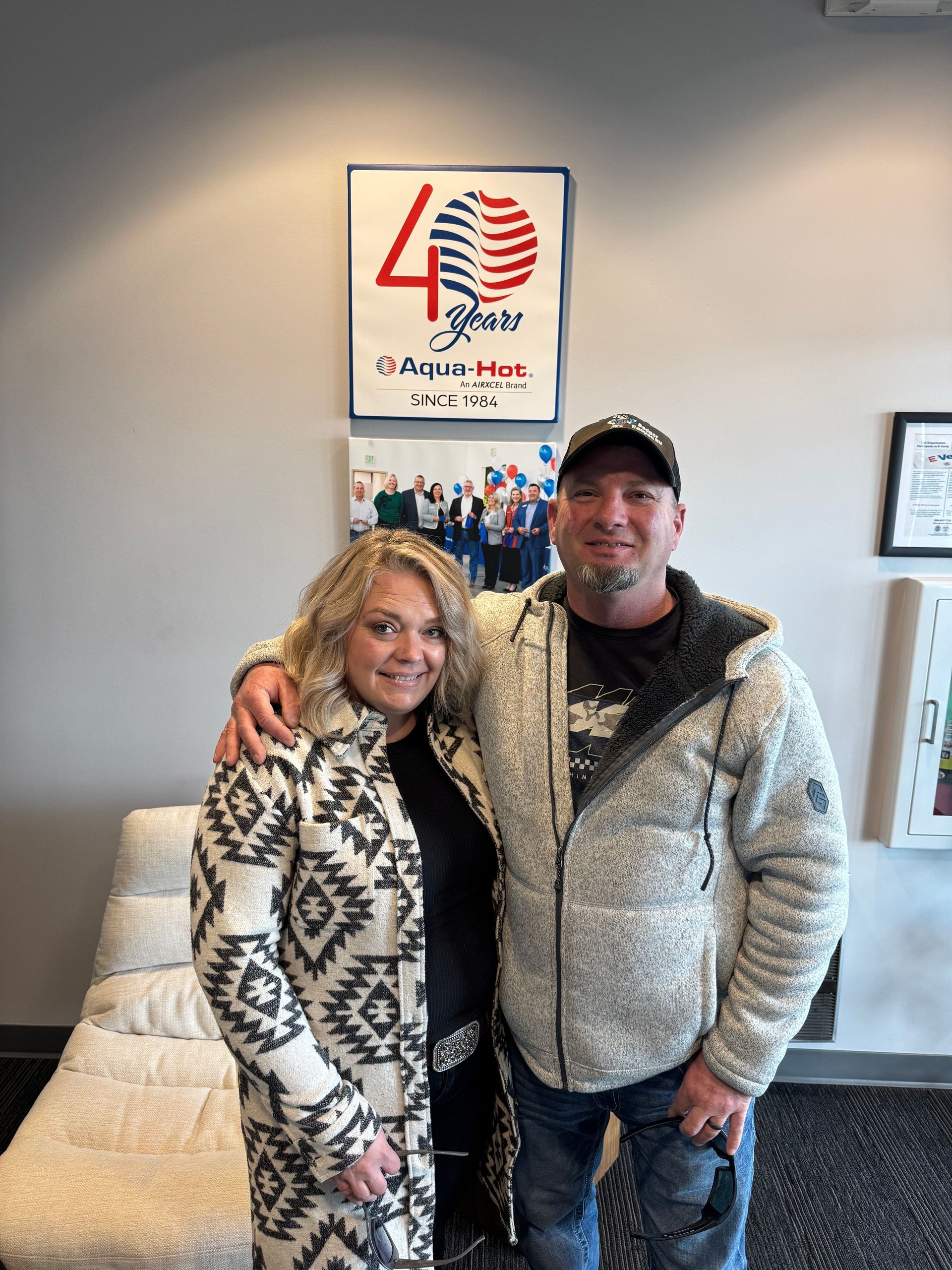 A couple poses in front of a 40-year anniversary flag display. The woman wears a patterned coat; the man a hooded jacket.