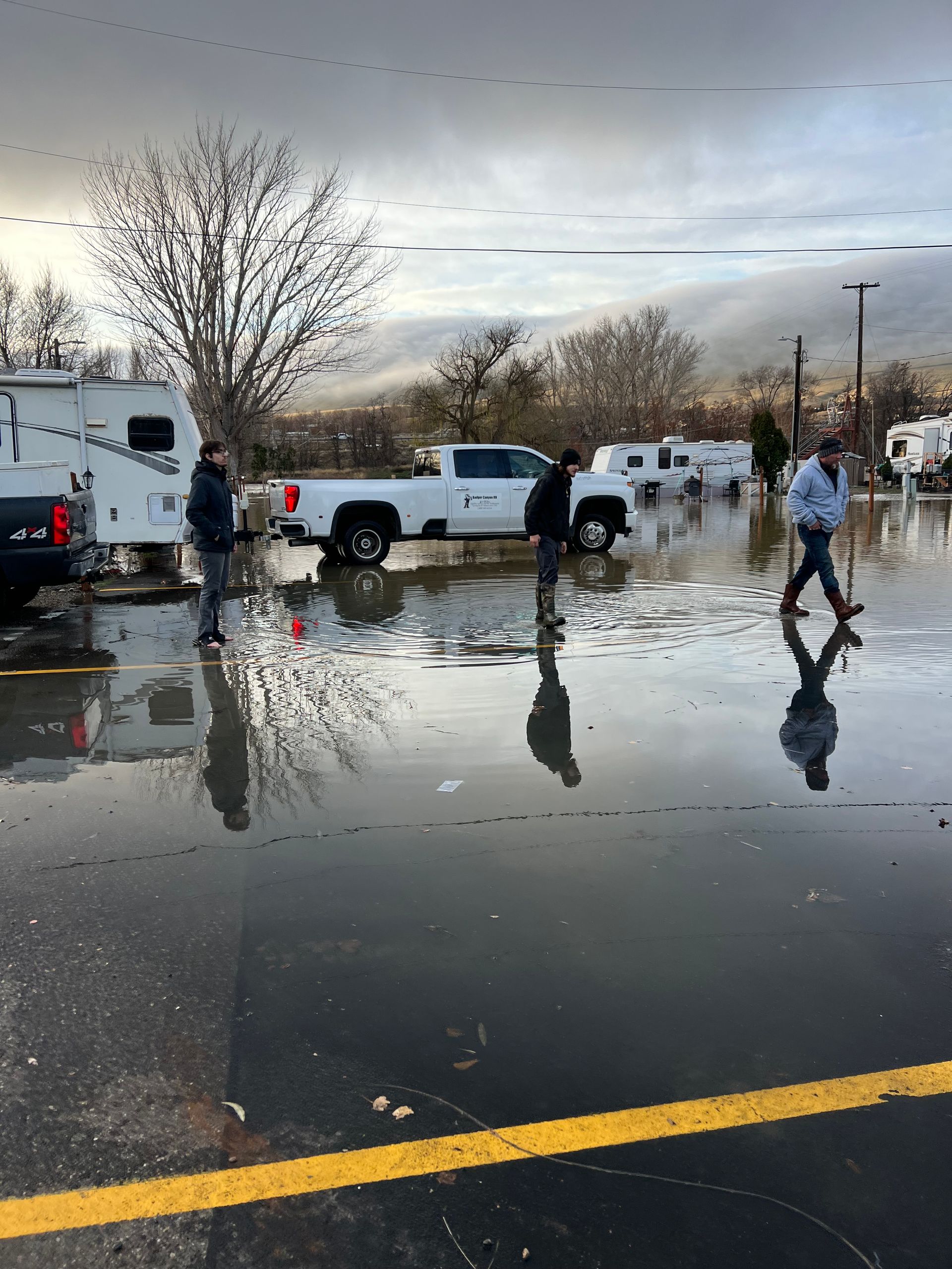 Flooded parking lot with people, pickup truck, and RVs. Cloudy sky, water reflection.