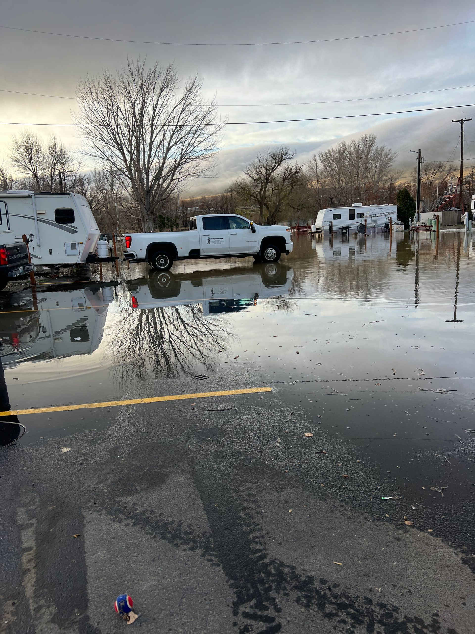 Flooded parking lot with a white pickup truck and several RVs, trees, and cloudy sky.