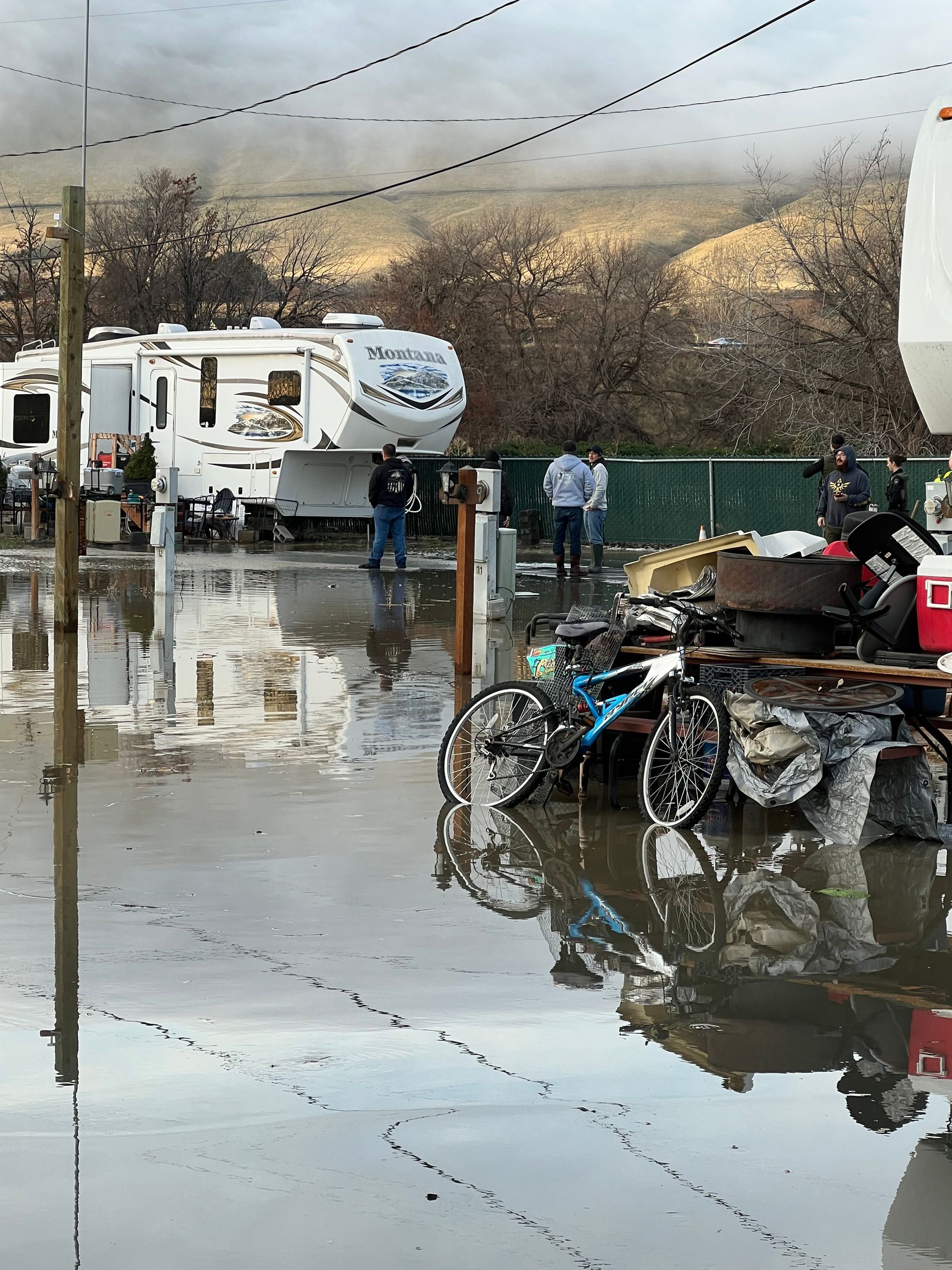 Flooded campsite with RVs. Two bicycles, people standing, waterlogged ground, overcast sky.