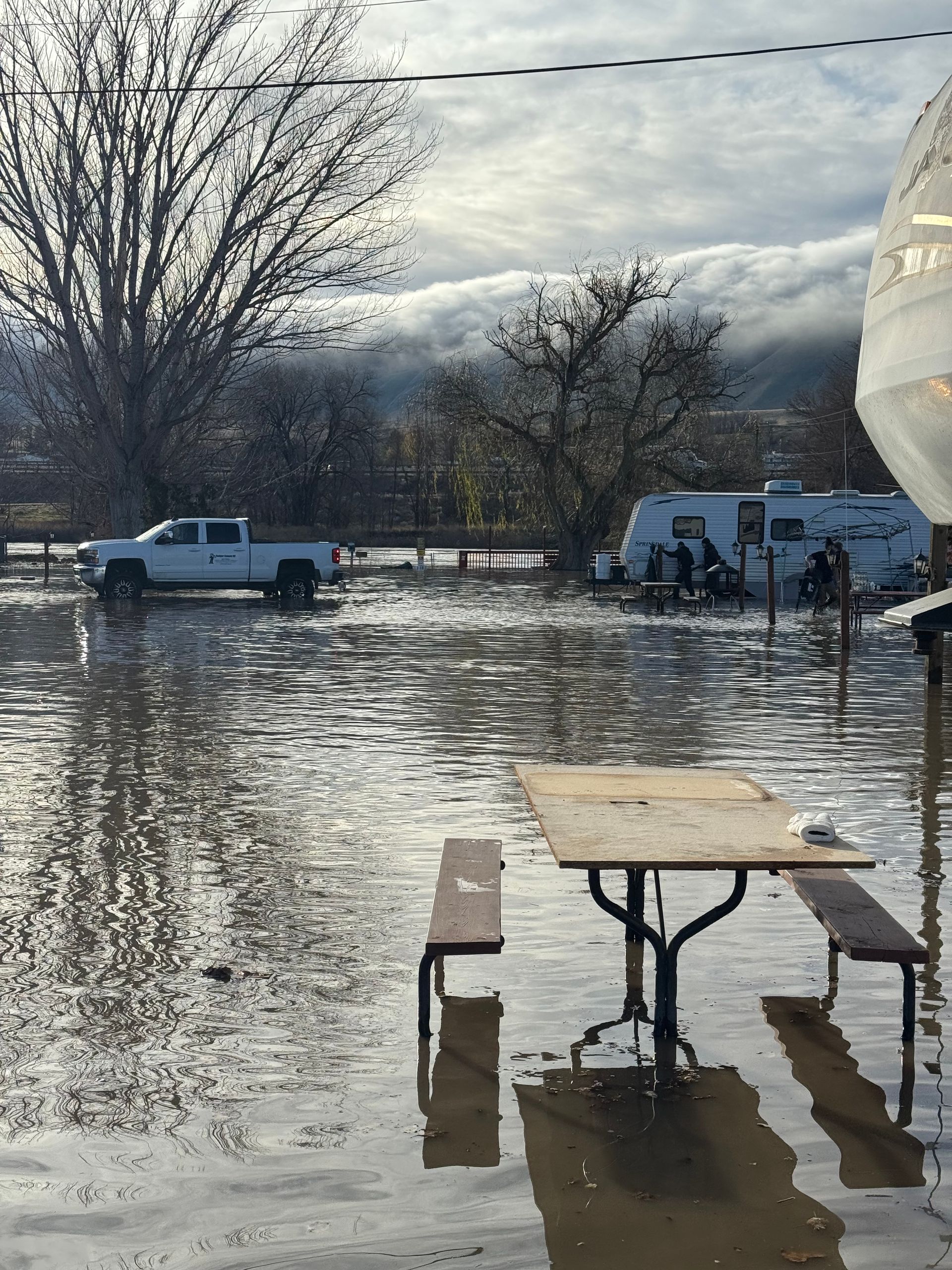 Flooded campsite: Picnic table submerged, truck and RV in water, overcast sky.